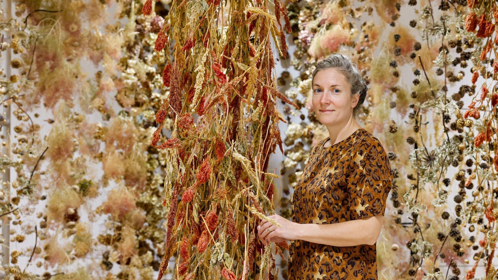 Artist Rebecca Louise Law standing beside red and yellow flowers suspended from the ceiling, Her grey hair is pulled back and she is wearing an orange top. She has blue eyes and is smiling slightly at the camera.