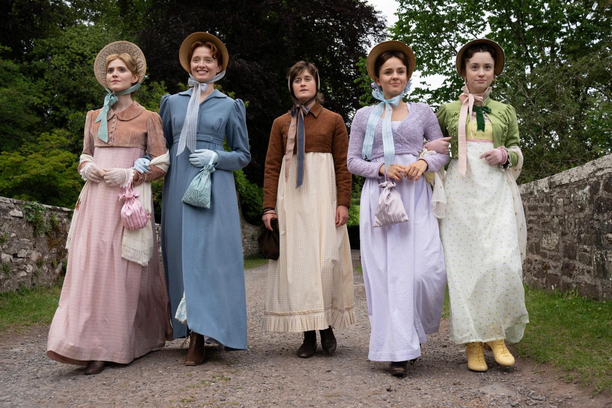 still from the other bennet sister. a full length shot showing the five bennet sisters walking arm in arm on a country lane. Maddie Close as Jane Bennet wears a pink dress, Poppy Gilbert as Lizzie Bennet is in a blue dress. Ella Bruccoleri as Mary Bennet wears a brown jacket with a yellow gingham skirt. Molly Wright as Kitty Bennet wears a purple dress and Grace Hogg-Robinson as Lydia Bennet wears a white dress with a small yellow floral print and a green jacket. everyone except mary bennet wears a bonnet, and mary wears a headscarf