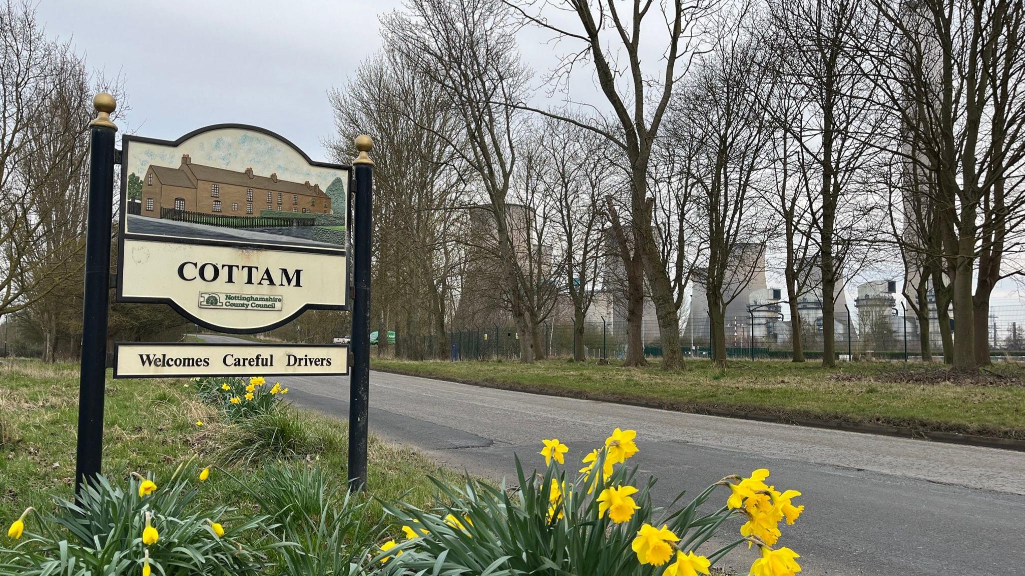 A village welcome sign with a power plant looming in the background