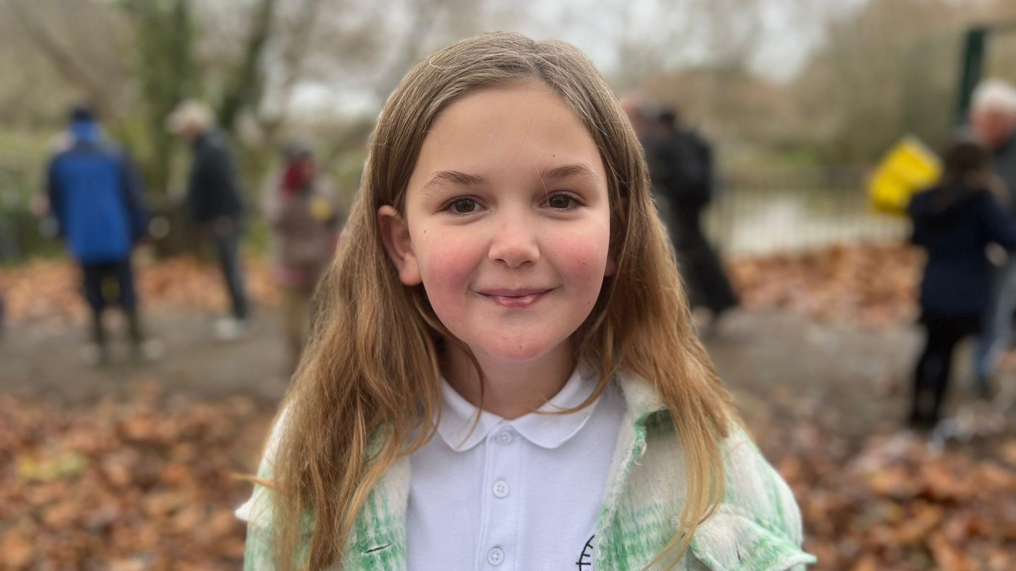 Darcie is smiling at the camera. She is standing outside in Castle Park with people gathered behind her. She is wearing a white school uniform polo shirt and a green and white checked jacket over the top