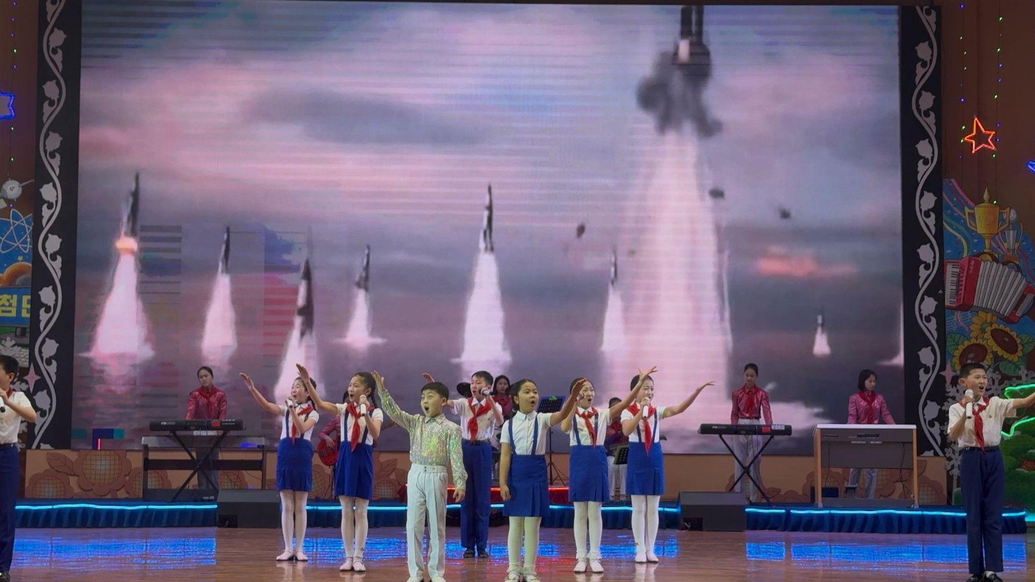 Young children stand on stage in front of a large screen with animations of ballistic missiles