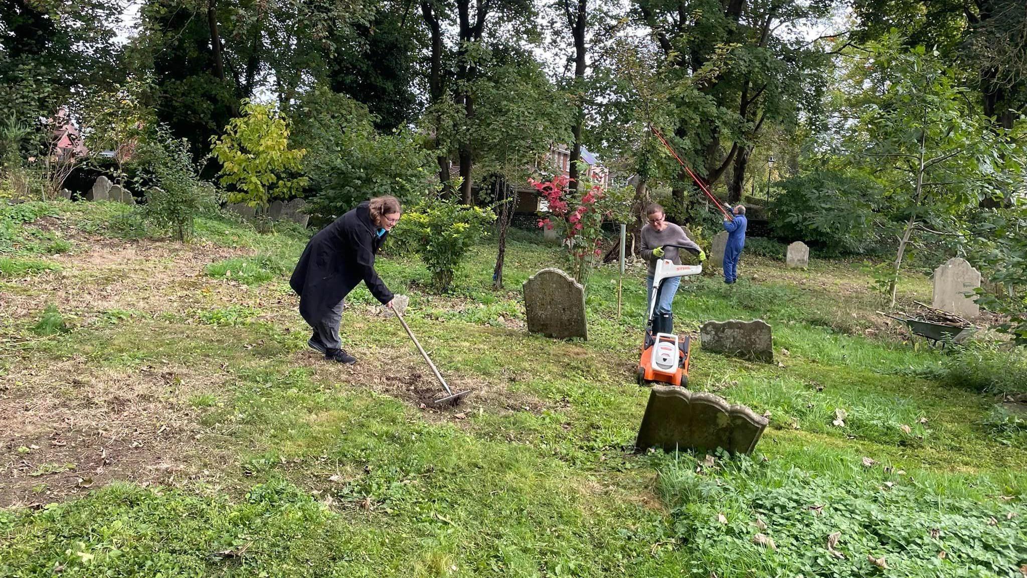 Two female volunteers rake and mow the grounds of the graveyard. Several gravestones are dotted about the site and tall trees are visible in the background.