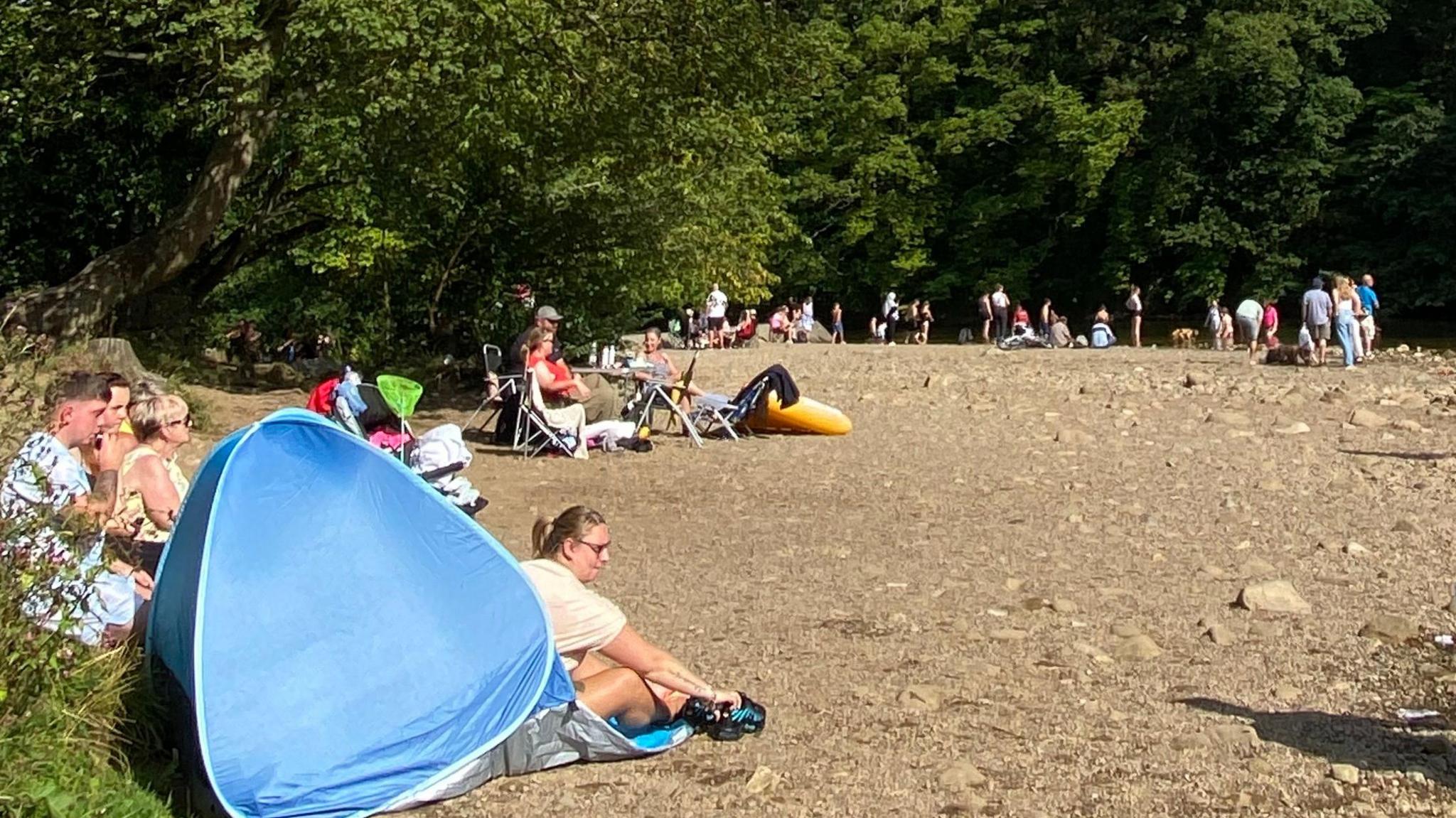People sit in tents and deck chairs on the bank of a river on a sunny day.