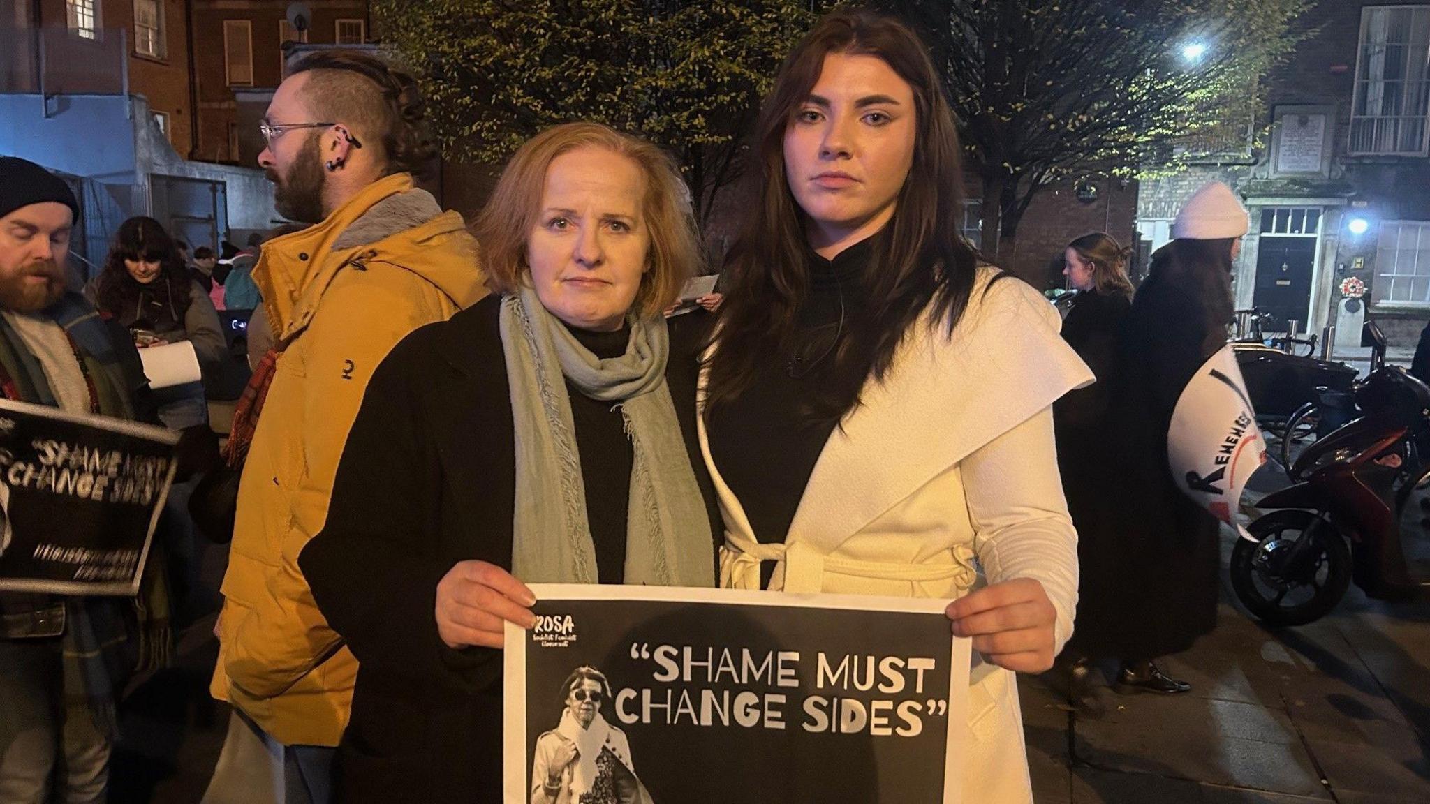 Ruth Coppinger in black coat and grey scarf and Natasha O'Brien in white coat (right)  - stood at rally, crowd visible in background includes a man in a bright yellow jacket