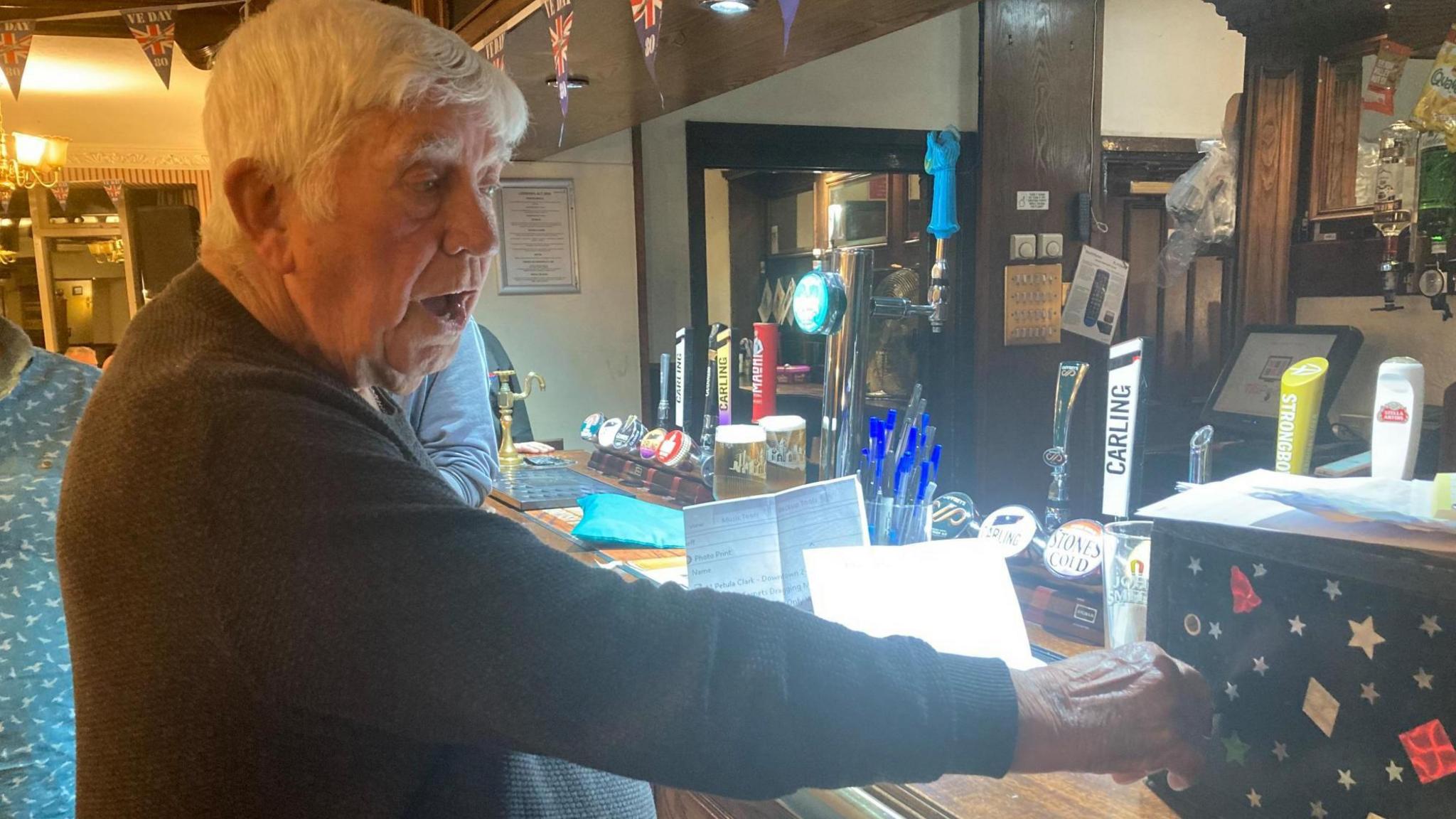An elderly man with white hair turns a key in the lock of a small black box on a bar