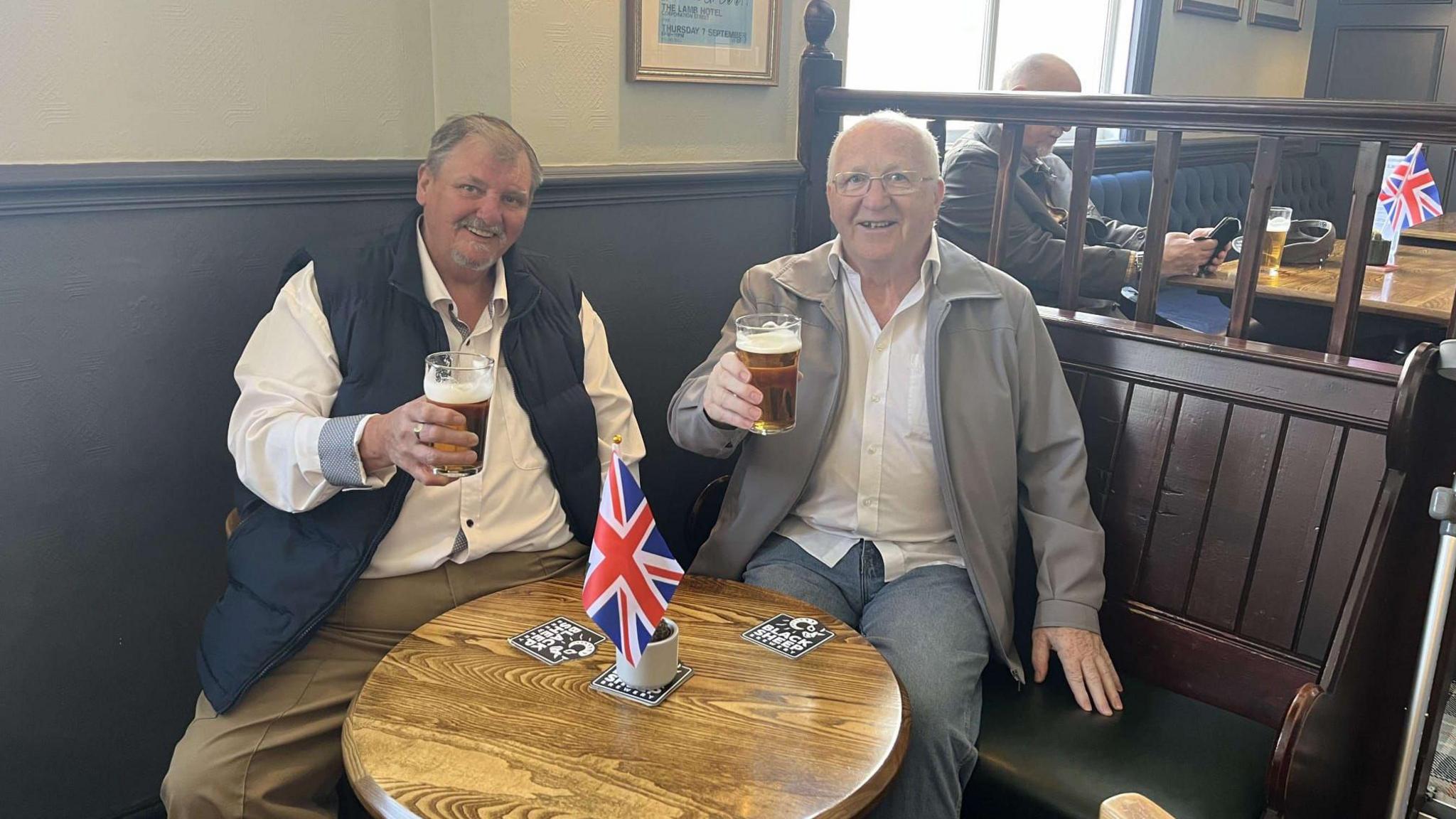 Two men sit at a pub table with union flags around them. They are smiling as they each hold a partly drink pint of beer.