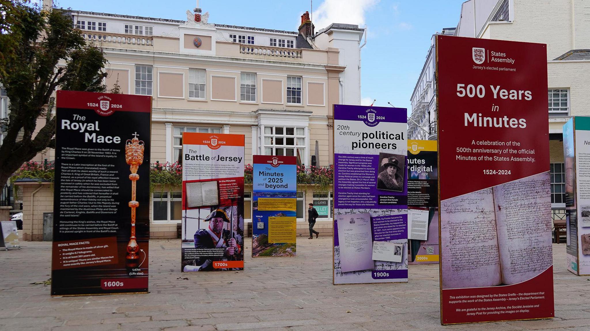 Six of the exhibition boards in the square, with titles including The Royal Mace, political pioneers and the Battle of Jersey. Buildings are visible in the background and to the side and a man walks in the gap between two boards in the distance.