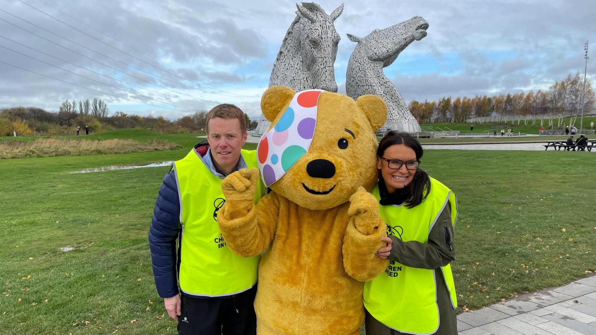 A man in a neon yellow bib is stood next to a person dressed as a yellow bear with a colourful eyepatch. A women also in a neon yellow bib is stood on the other side of the bear. The trio are stood in front of two metal horse heads on a patch of grass.