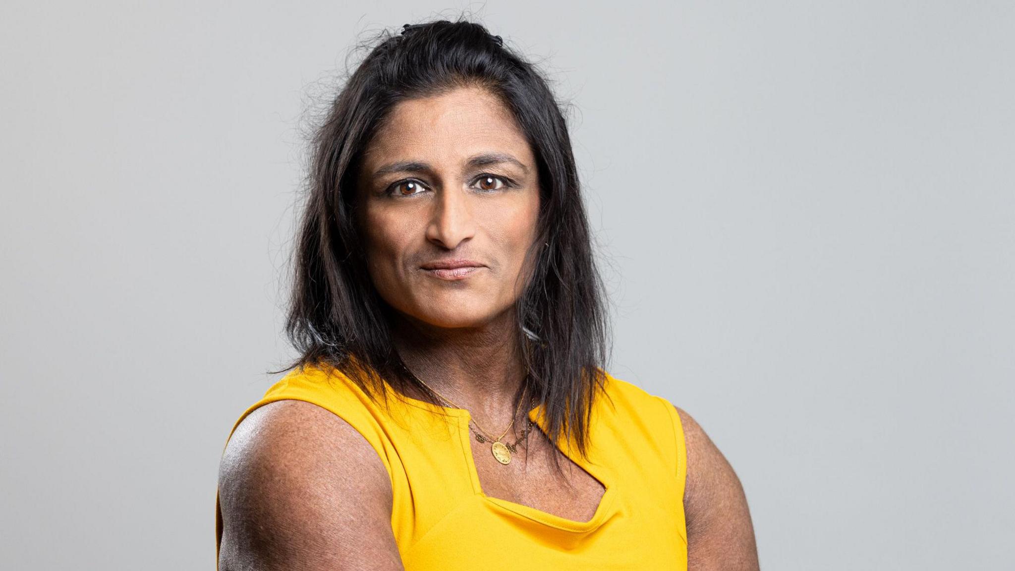 Ajanta Hilton with long dark hair smiling at the camera while wearing a yellow T-shirt