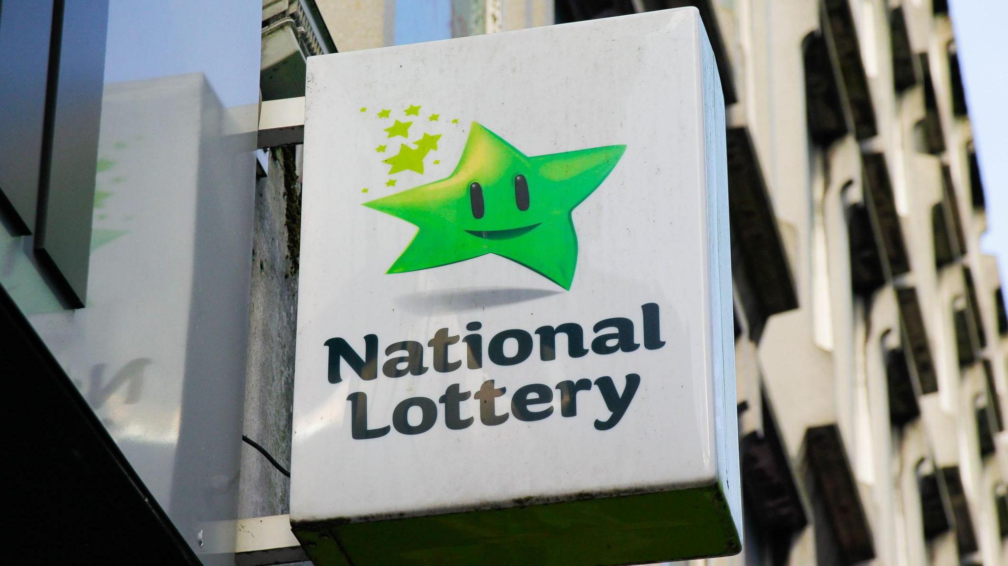 A white exterior sign hangs from a grey wall. The sign has a green star, with a black mouth and black eyes. Below the star are the words "National Lottery."