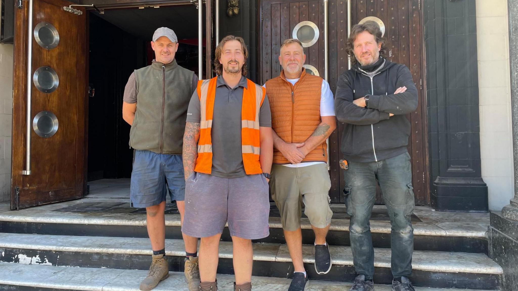 Four men in battered work gear stand proudly outside the steps to an entrance of a large building
