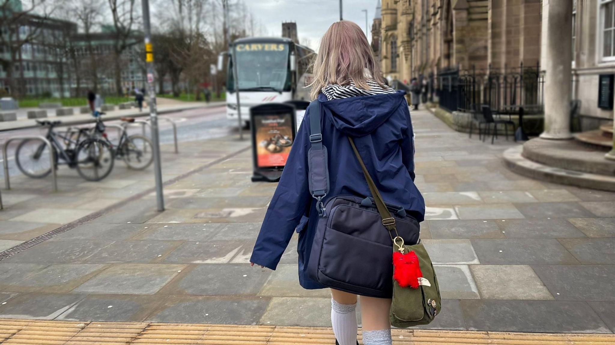 Amelka wearing a navy rain jacket and two bags on her back is walking towards her university campus. A bus can be seen on the road and a bike rack nearby with two bicycles parked up outside the campus.