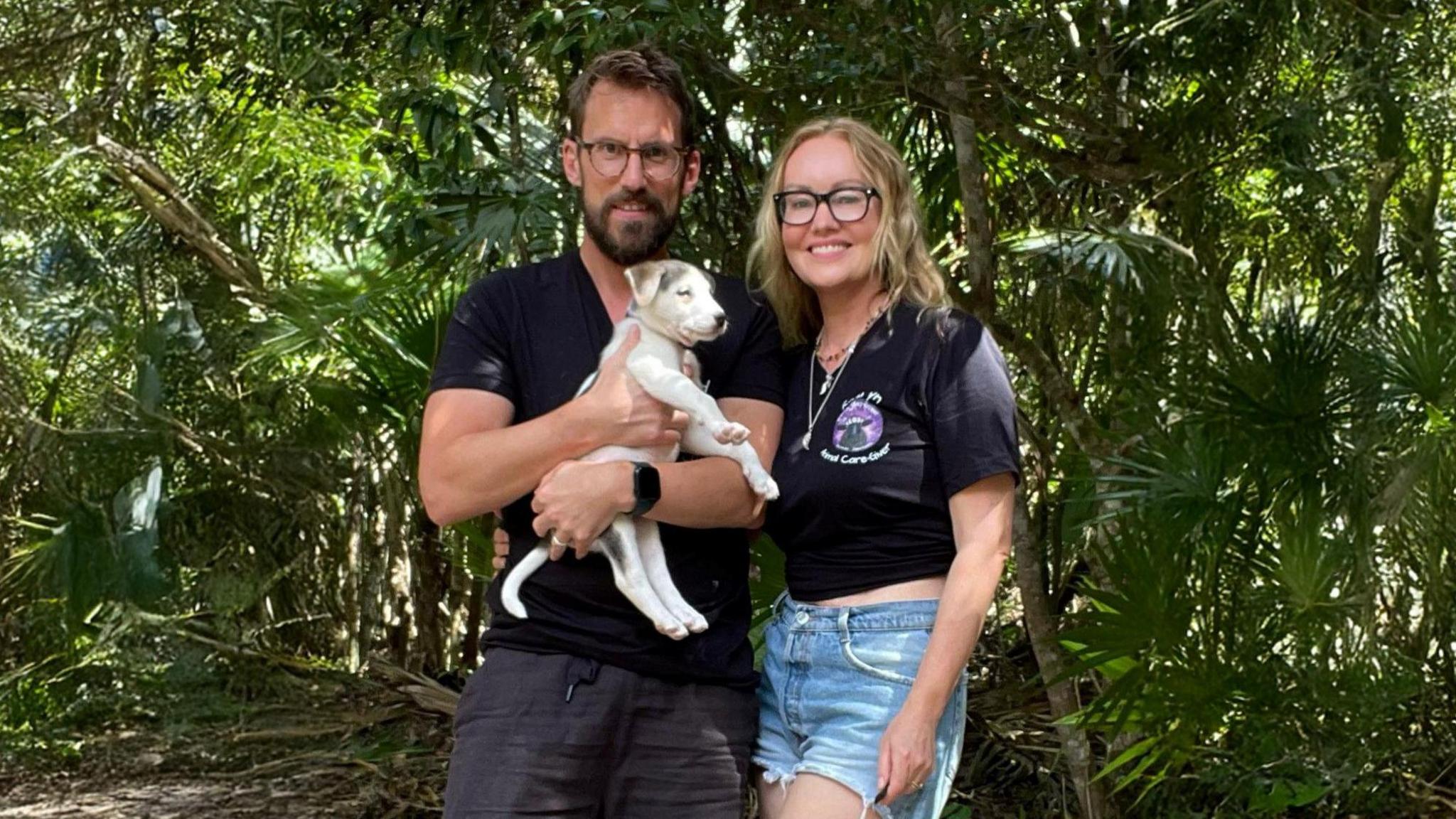 David Brudnell is holding a small light-coloured puppy in his arms. He has a dark T-shirt on and has short dark hair and a beard. He is standing next to Kathryn Grace who has long blonde hair and is wearing a T-shirt and shorts. There are trees behind them