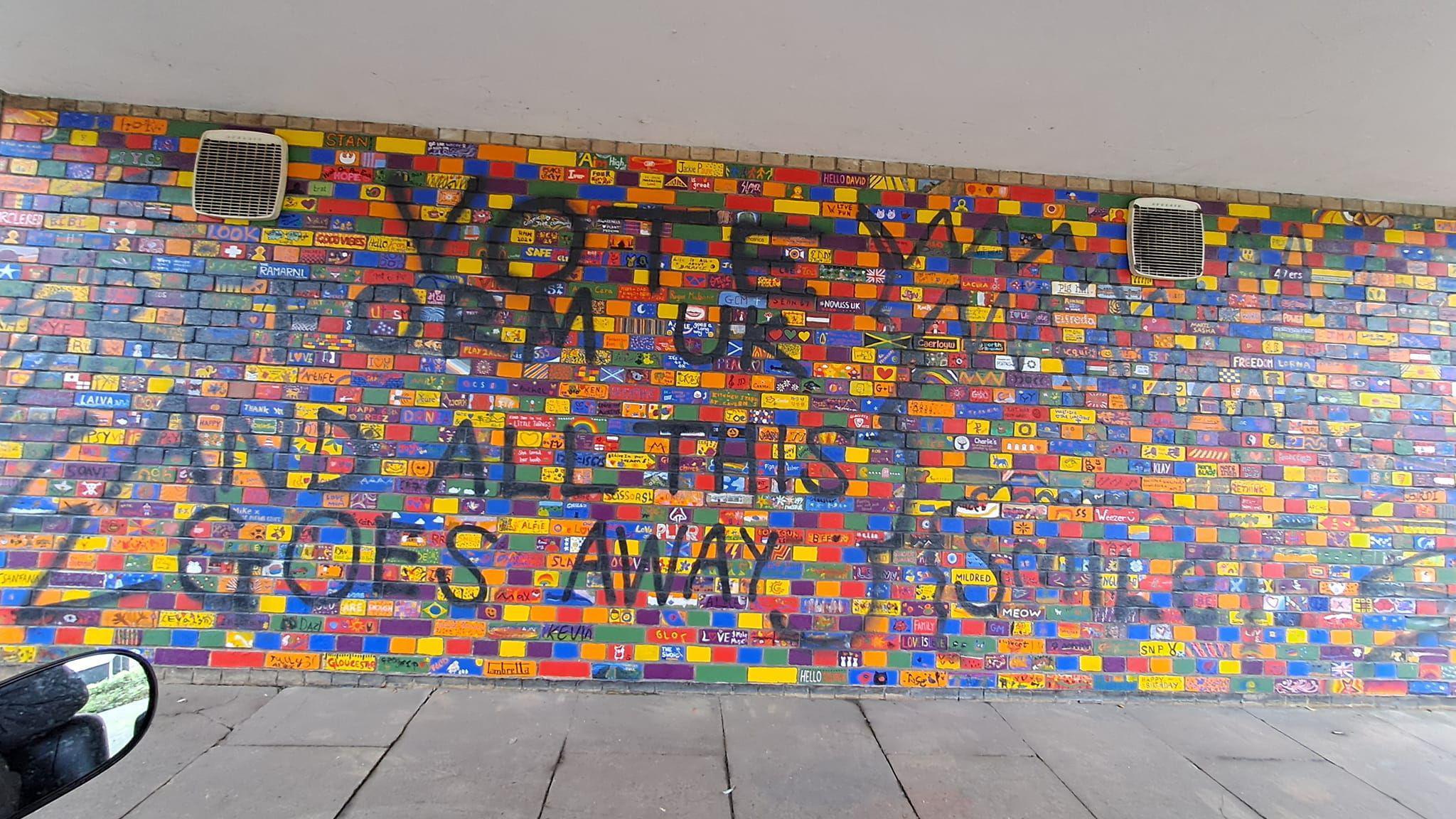 A brick wall in a tunnel. Around 1,500 bricks are individually painted bright colours, some with symbols and flags, others with animals and local landmarks. Over the top of it someone has spray painted the phrase "Vote Reform UK and this all goes away" 