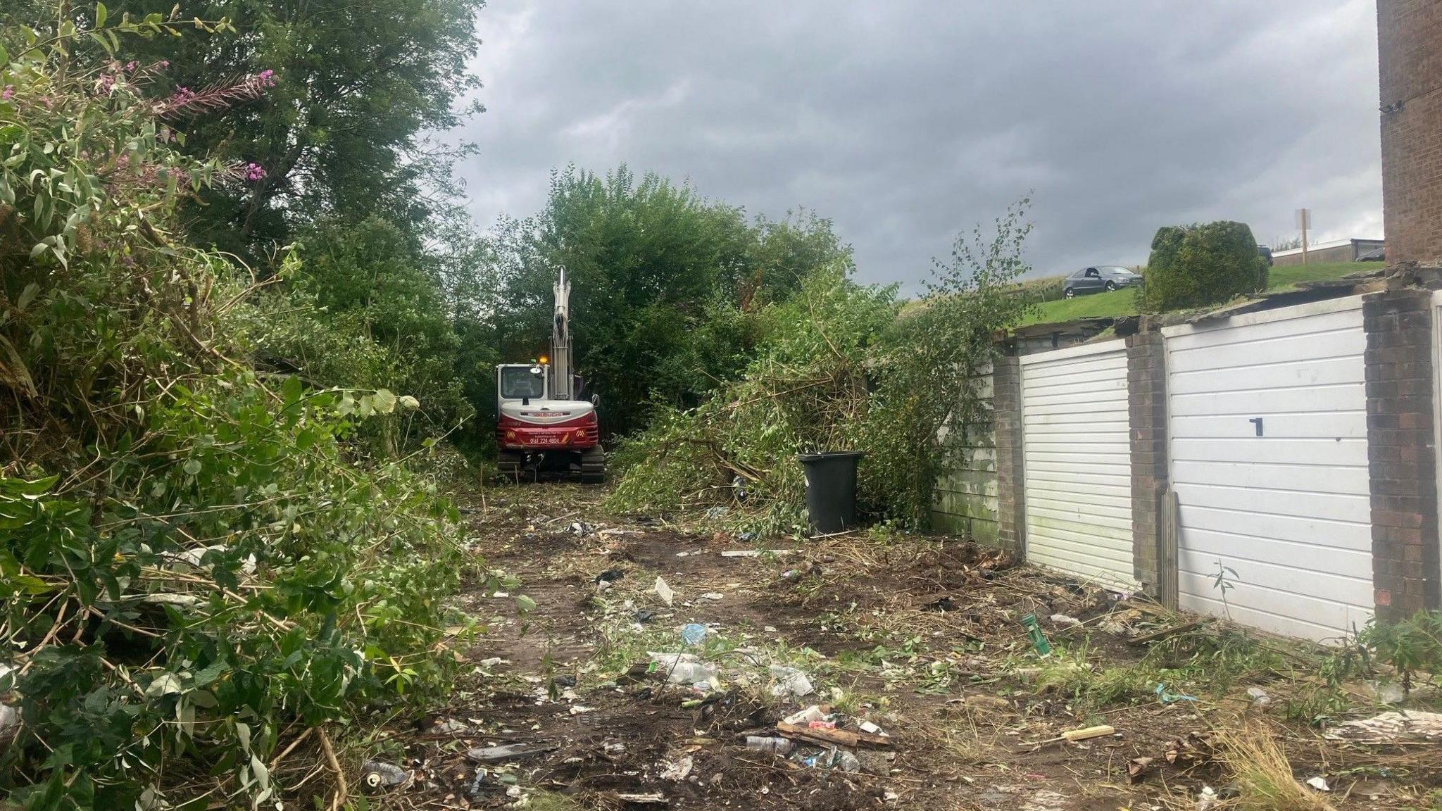 Photo of run-down garages to the right of the image with a digger and bushes to the left