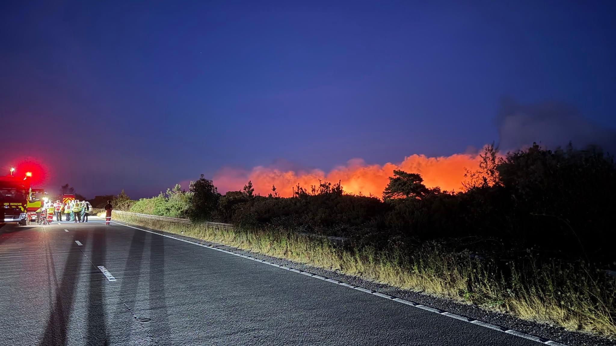 Fire in New Forest with orange glow seen to the right of a road and many emergency services in the distance.