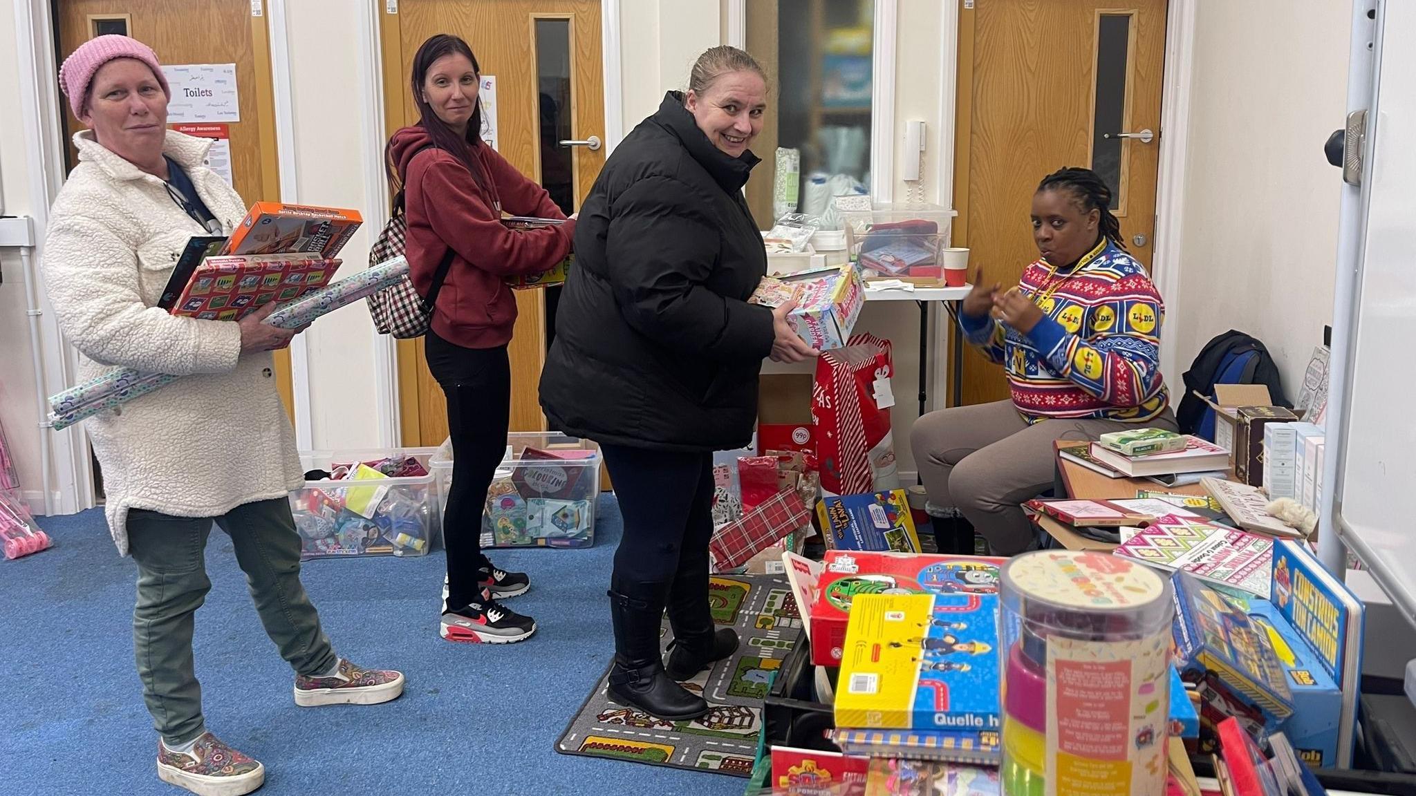 Three women lining up holding presents surrounded by a range of games, books and wrapping accessories. 