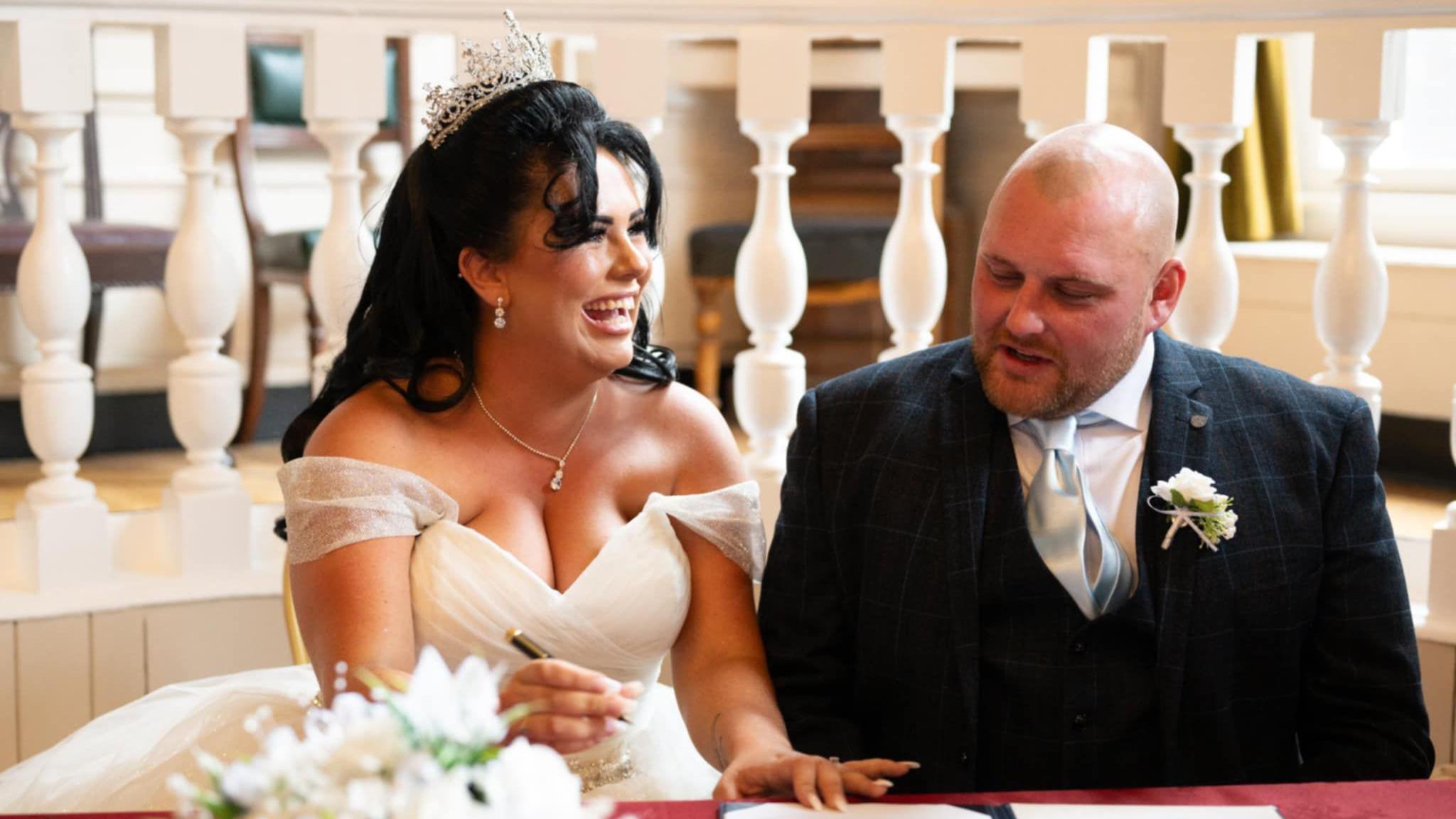 Shania Stanton and her husband on their wedding day signing the marriage schedule. She is on the left wearing a white dress with exposed shoulders and a tiara. She has long dark hair and is laughing and holding the pen. He is wearing a suit with a blue silk tie and a flower brooch. He is bald. There is a bouquet in the foreground. 