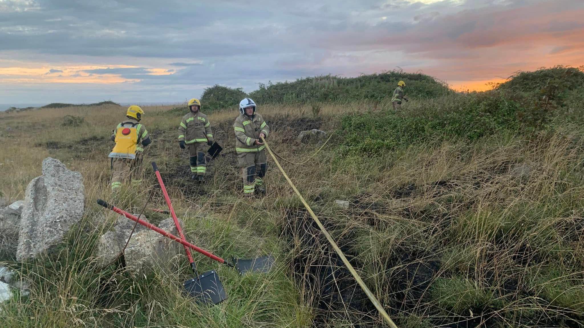 Three firefighters wearing uniform overalls and helmets working in an area of grassland. In the foreground is a charred area of ground. Two beaters are propped up against a large rock. One of the firefighters is pulling a yellow hose. The other end is being held by a fourth firefighter standing in the distance.