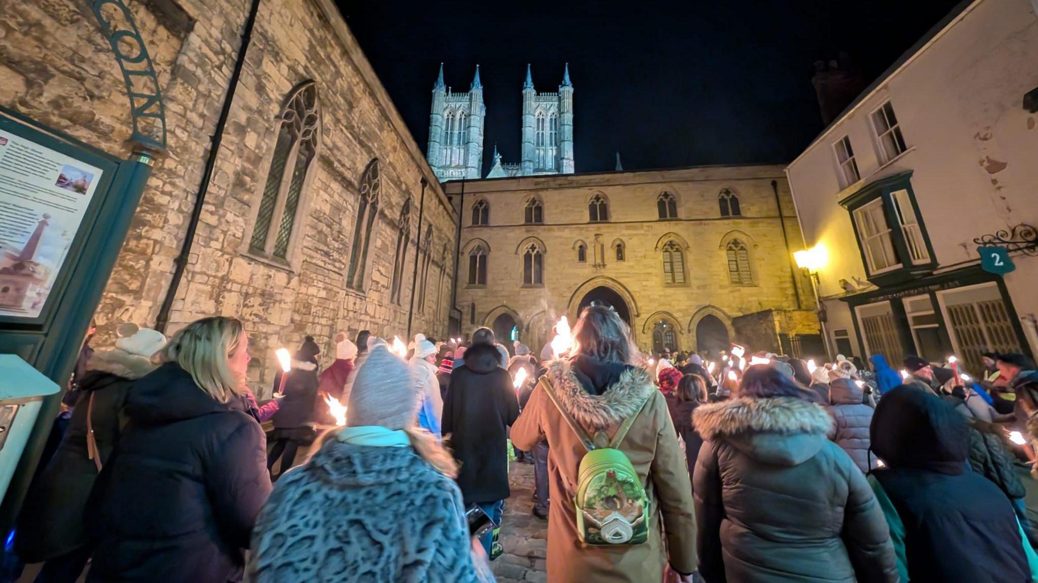 The memorial parade makes its way through Castle Square in Lincoln towards the city's cathedral which is visible in the background. The participants are holding long candles which have burning flames. The group is surrounded by historic stone buildings which are illuminated by lights and the walkers. The dark night sky is overhead.