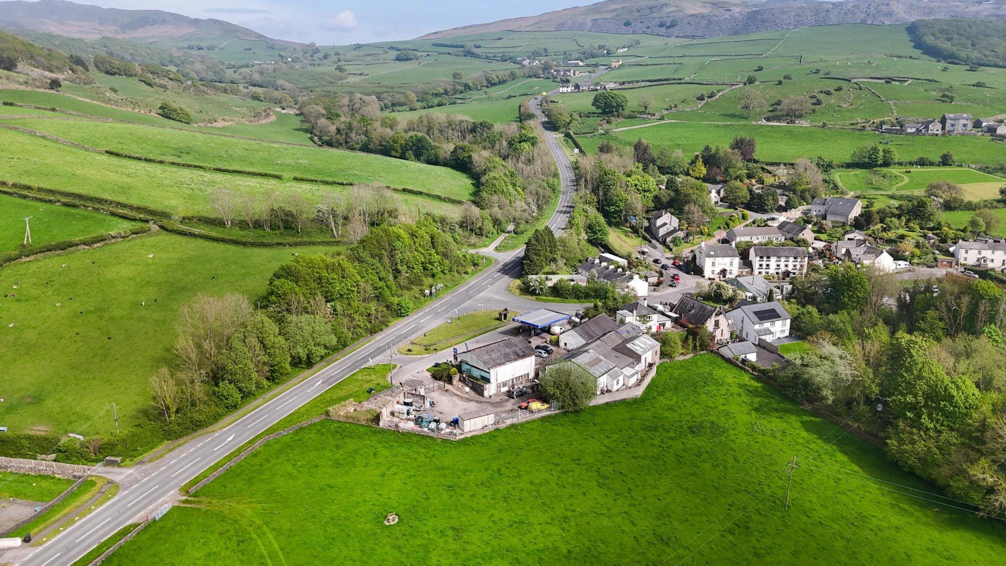 Aerial view of the area showing an existing road, surrounding fields and a number of white houses and other buildings.