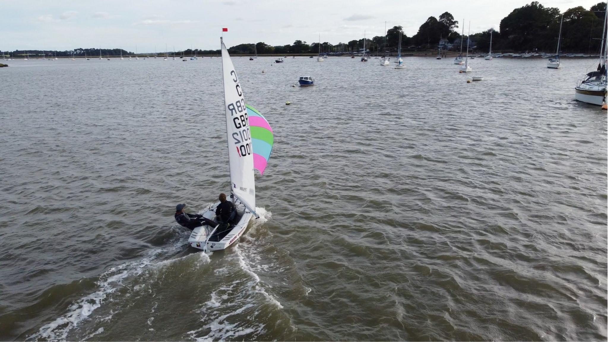 Alex and Jude sail together on the River Deben in Suffolk in the late evening sunshine. They're in the middle of the river with boats and Waldringfield's clubhouse to the right and other boats ahead further down the river.