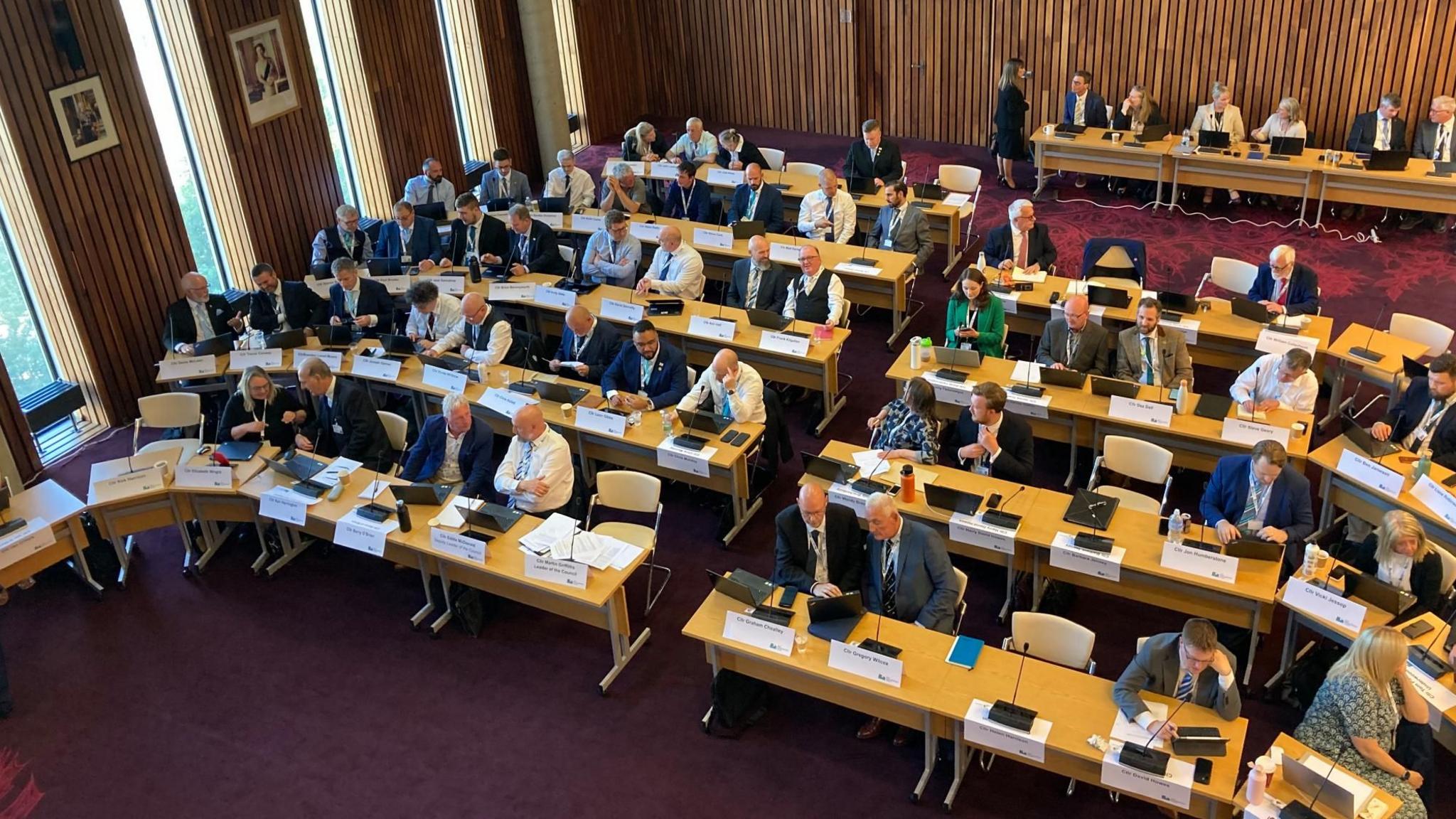 A bird's eye view of about 60 councillors sitting at five rows of tables in a horseshoe shape in a council chamber. The carpet is a deep red and the walls are wooden panelled. There is a separate row of tables at the back of the room with half a dozen people sitting there.