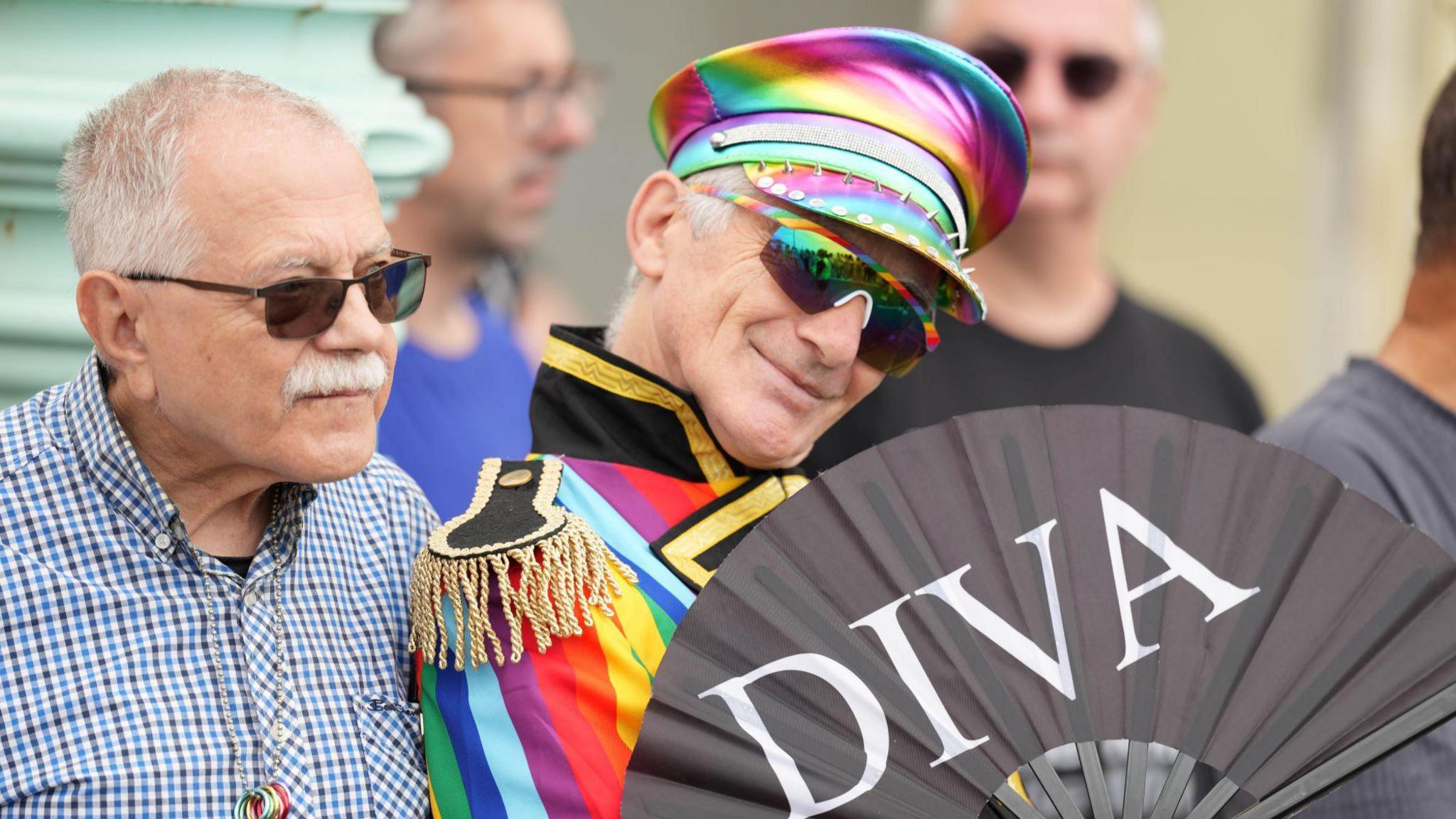 A man with a rainbow hat on holding a black fabric fan. The fan has white writing on reading "Diva".