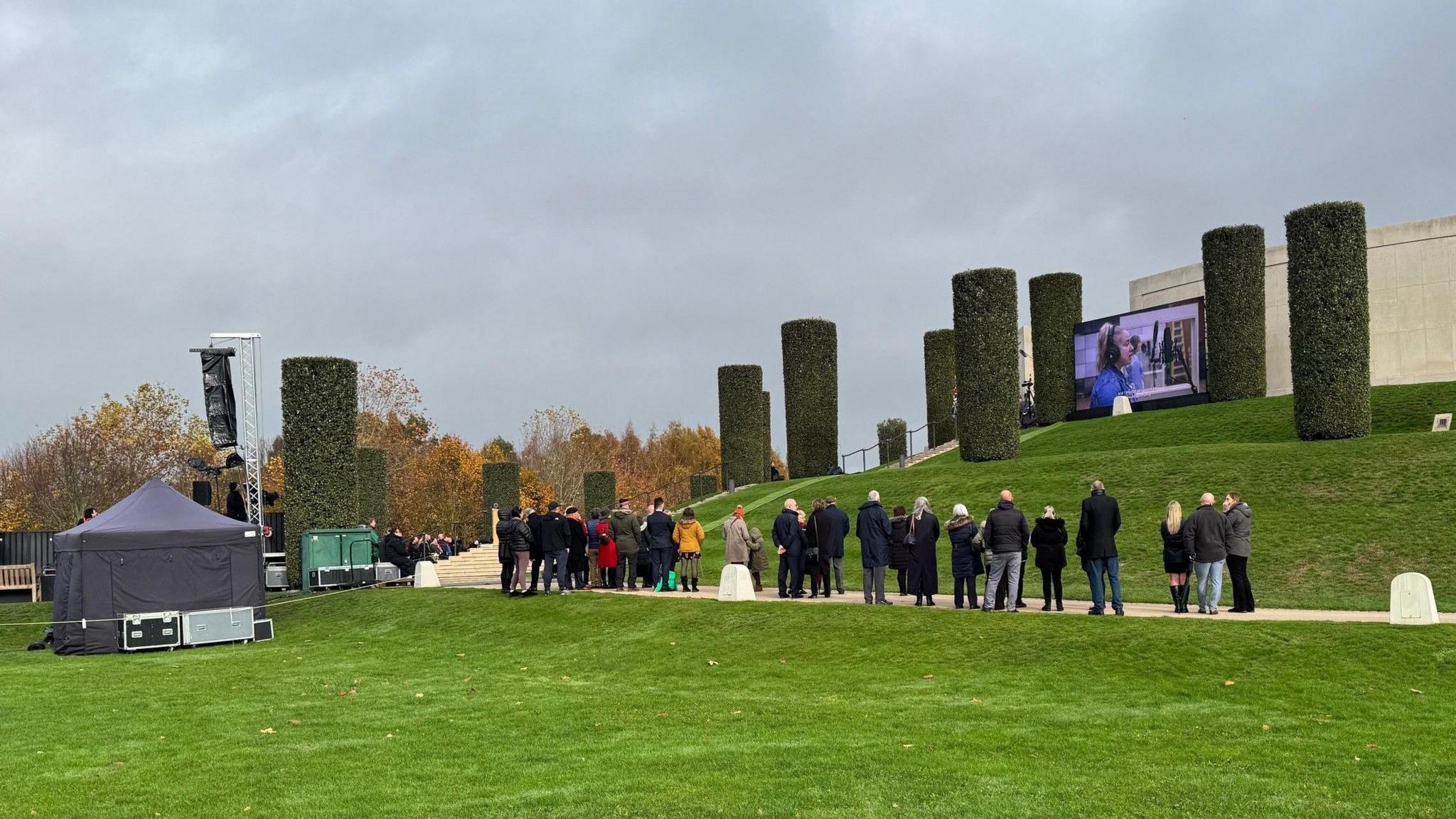 A row of people standing at the foot of a grassy hill with trees cut into the shape of pillars and a concrete structure at the top with a large TV screen in front of it