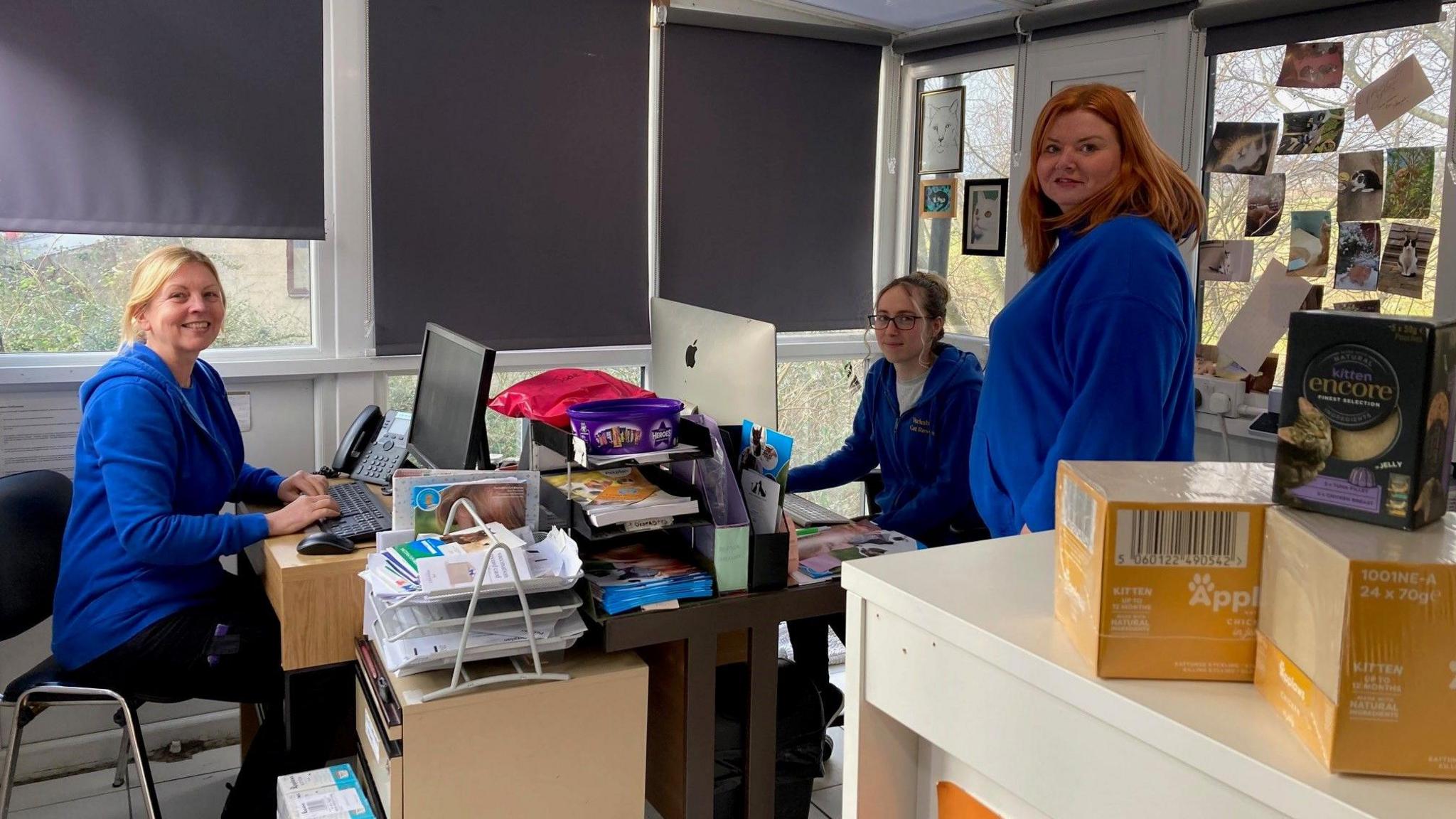 Three woman all wearing light blue, zip-up fleeces in an office with dual aspect, double-glazed windows.  