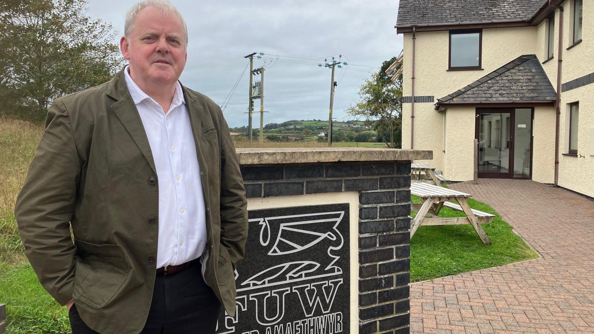 Guto Bebb yn sefyll o flaen arwydd Undeb Amaethwyr Cymru, tu allan i'r swyddfa. Mae'n edrych ar y camera ac yn gwisgo crys gwyn gyda siaced wyrdd a throwsus du. Mae ganddo wallt llwyd.