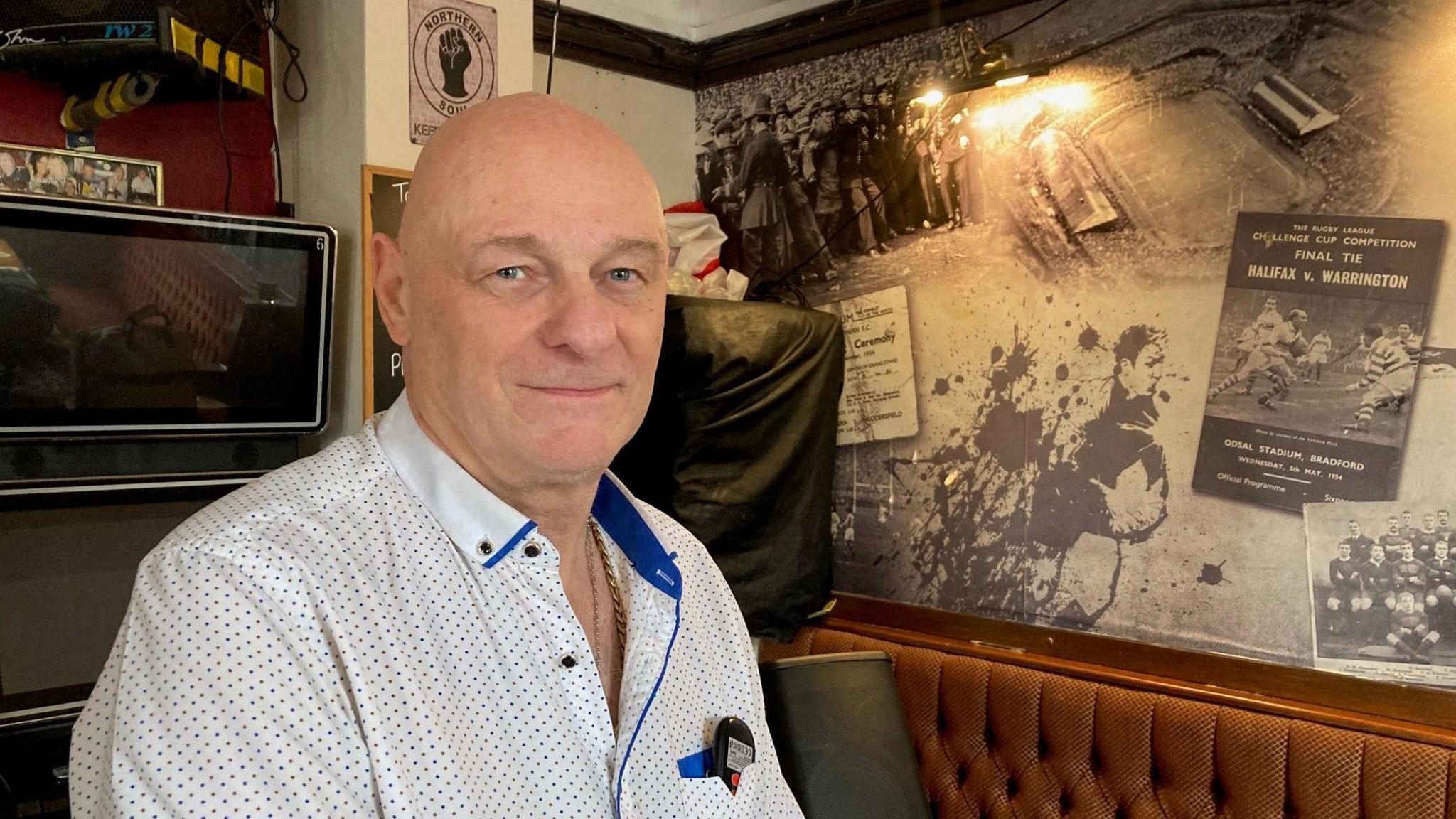 A bald man wearing a white shirt with a blue and brown dot pattern standing next to a wall with sporting posters on it.