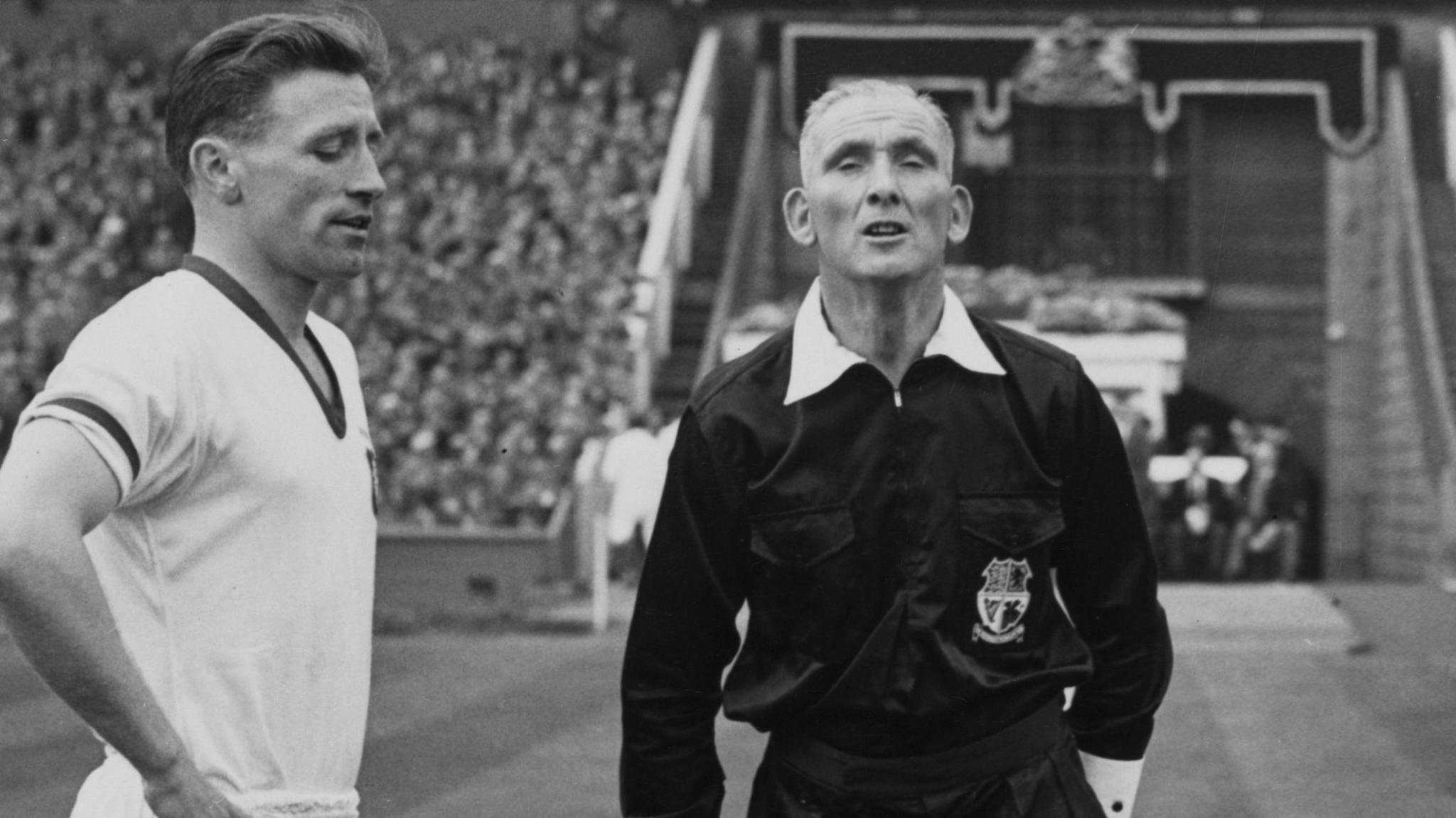 A black and white photo of two men from the waist up, one is wearing a white football shirt while the other is dressed in a referee's traditional black, long sleeved shirt . In the background there is a crowd of people sitting in a stadium