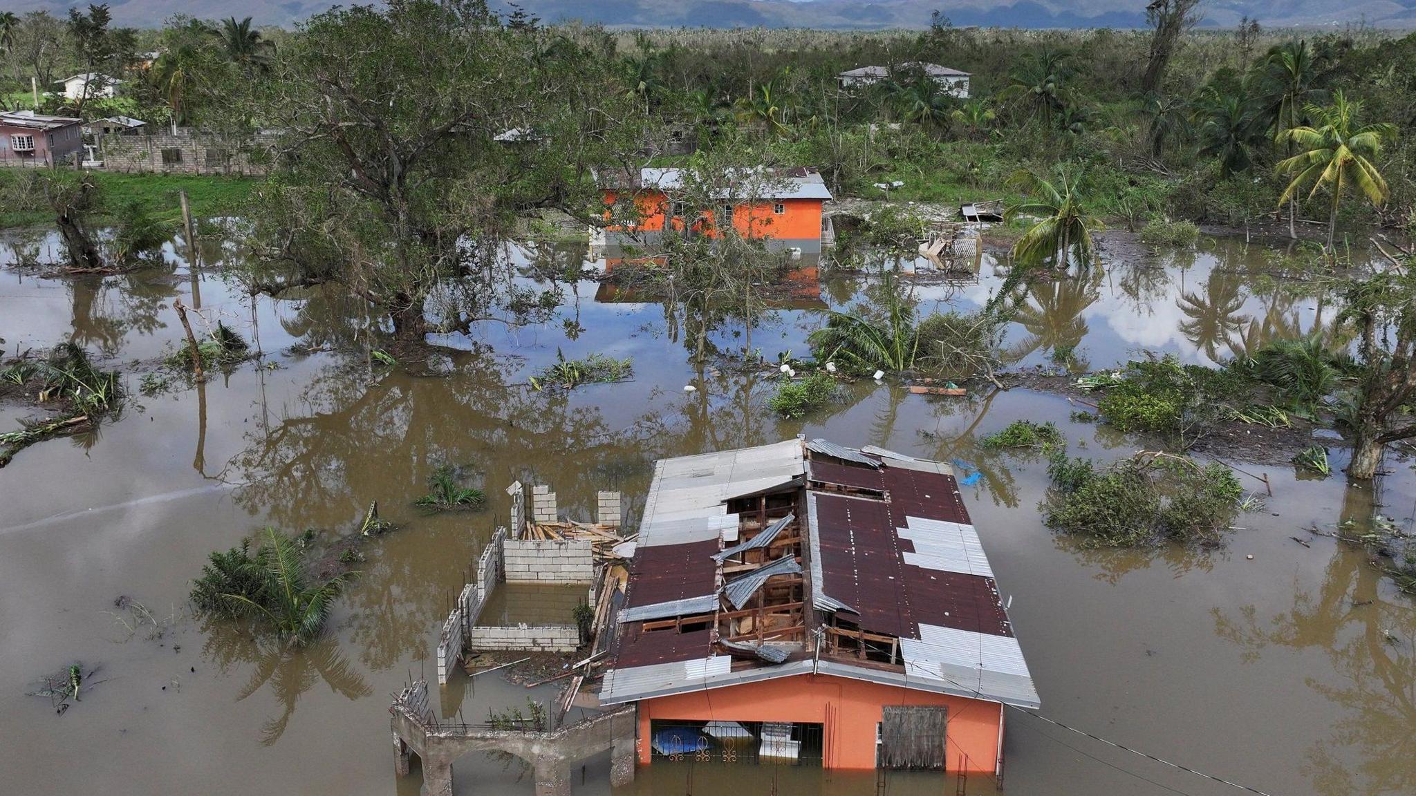 A drone image of a landscape that has been battered by a hurricane. It is flooded with green trees either standing or toppled over. There is a house in the foreground that is flooded, with pieces of the roof missing.