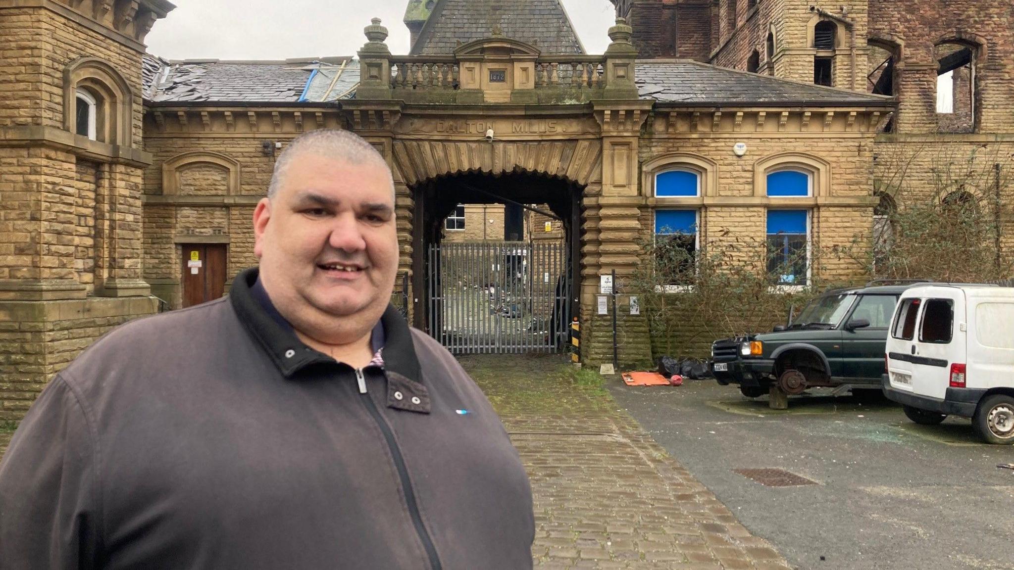 A man wearing a black jacket standing outside a honey coloured stone building with an ornate entrance featuring a pillar either side and other architectural features above.