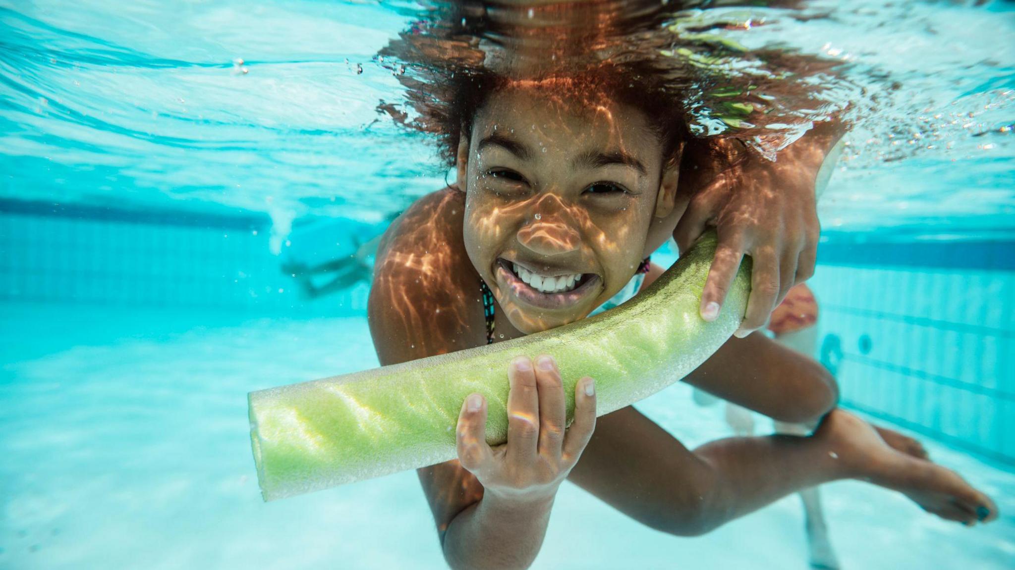 A child is smiling whilst holding a pool buoyancy aid underwater in a swimming pool.