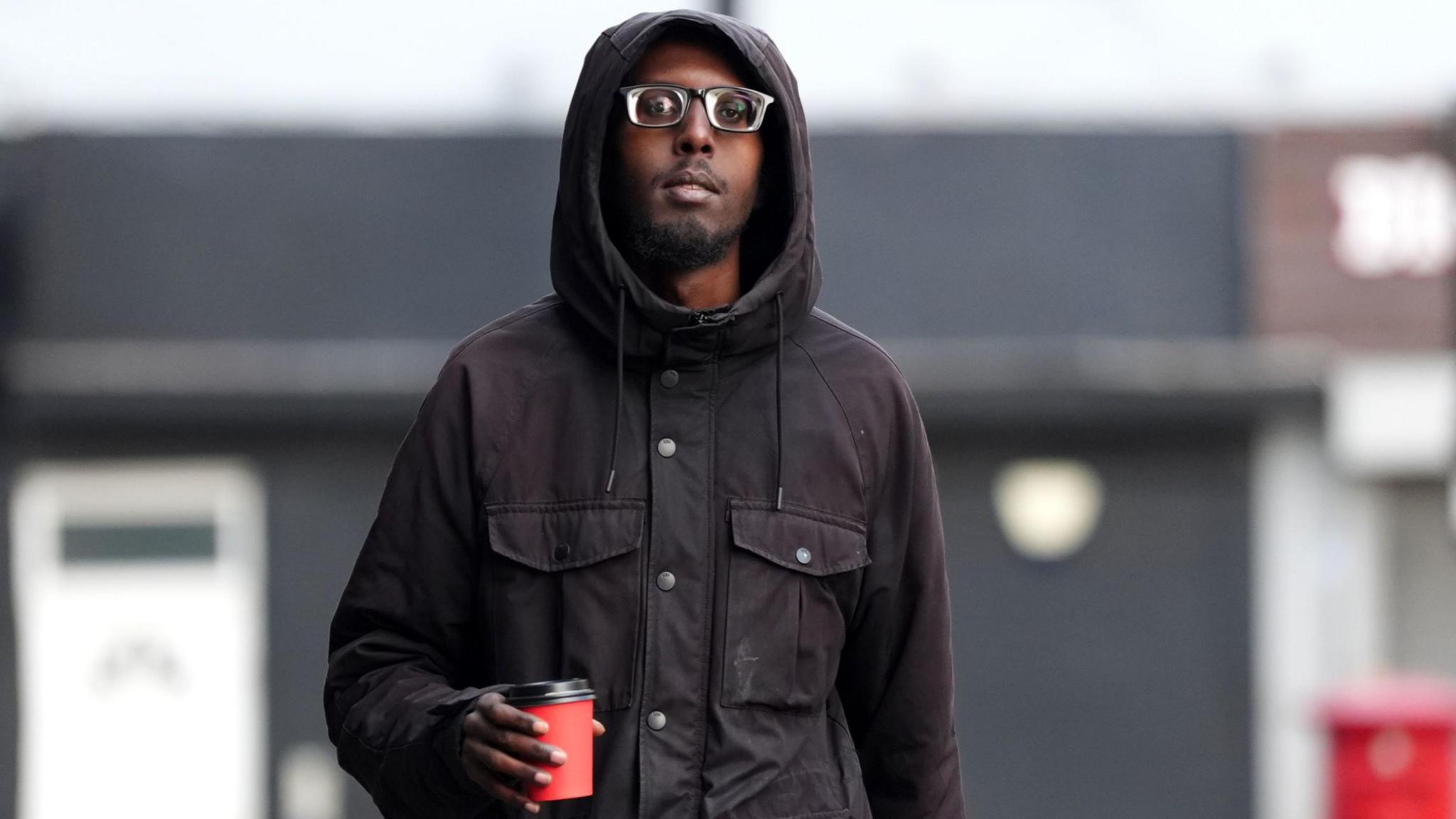 Ayub Dirie dressed in black trousers and black hooded jacket clutches a red coffee cup