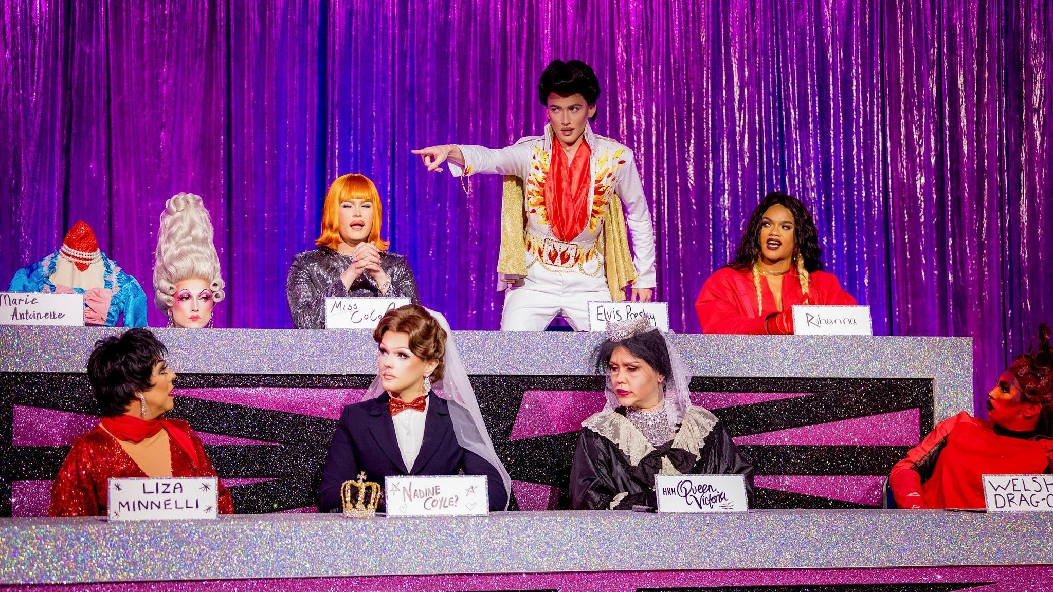 Contestants compete in the Snatch Game. Sitting in two rows from L-R: Marmalade is a beheaded Marie Antoinette; Chanel O'Conor is Coco Peru; Kyran, dressed as Elvis Presley, stands and points at RuPaul; Rileasa is Rihanna; La Voix looks at Kyran while dressed as Liza Minnelli; Charra Tea is Nadine Coyle dressed as Mary Poppins; Lill is Queen Victoria and Actavia is the Welsh dragon. 