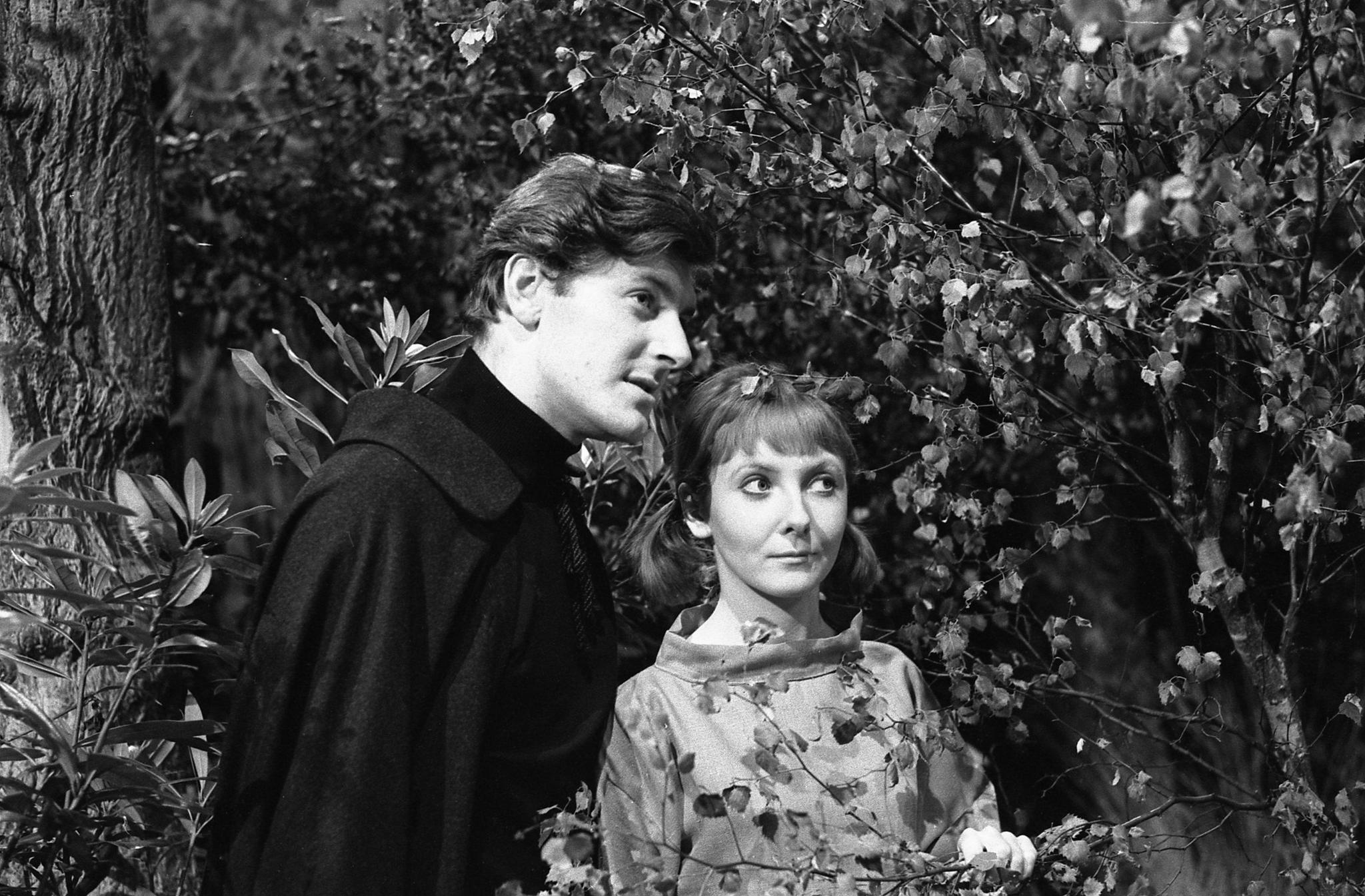 Black and white photo of Steven Taylor, who has short dark hair and is wearing a black cloak, and Vicki, who has lighter hair in short pigtails and is wearing a light coloured cowl-neck top, look out at something to their left. They are standing in amongst trees and bushes.