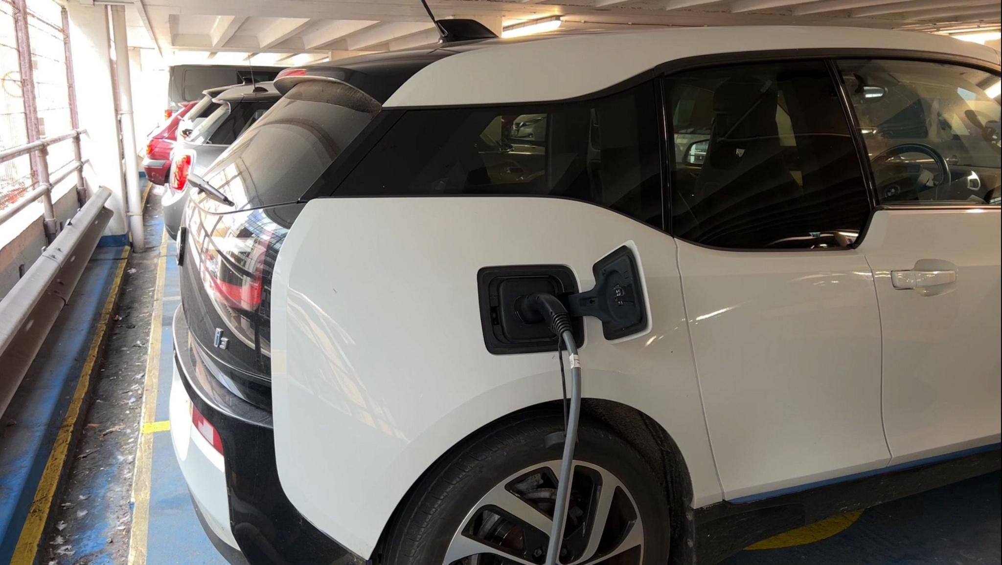 A white hatchback is reverse parked in an underground car park. It is being charged by an electric vehicle charger, which is connected to its charging point.