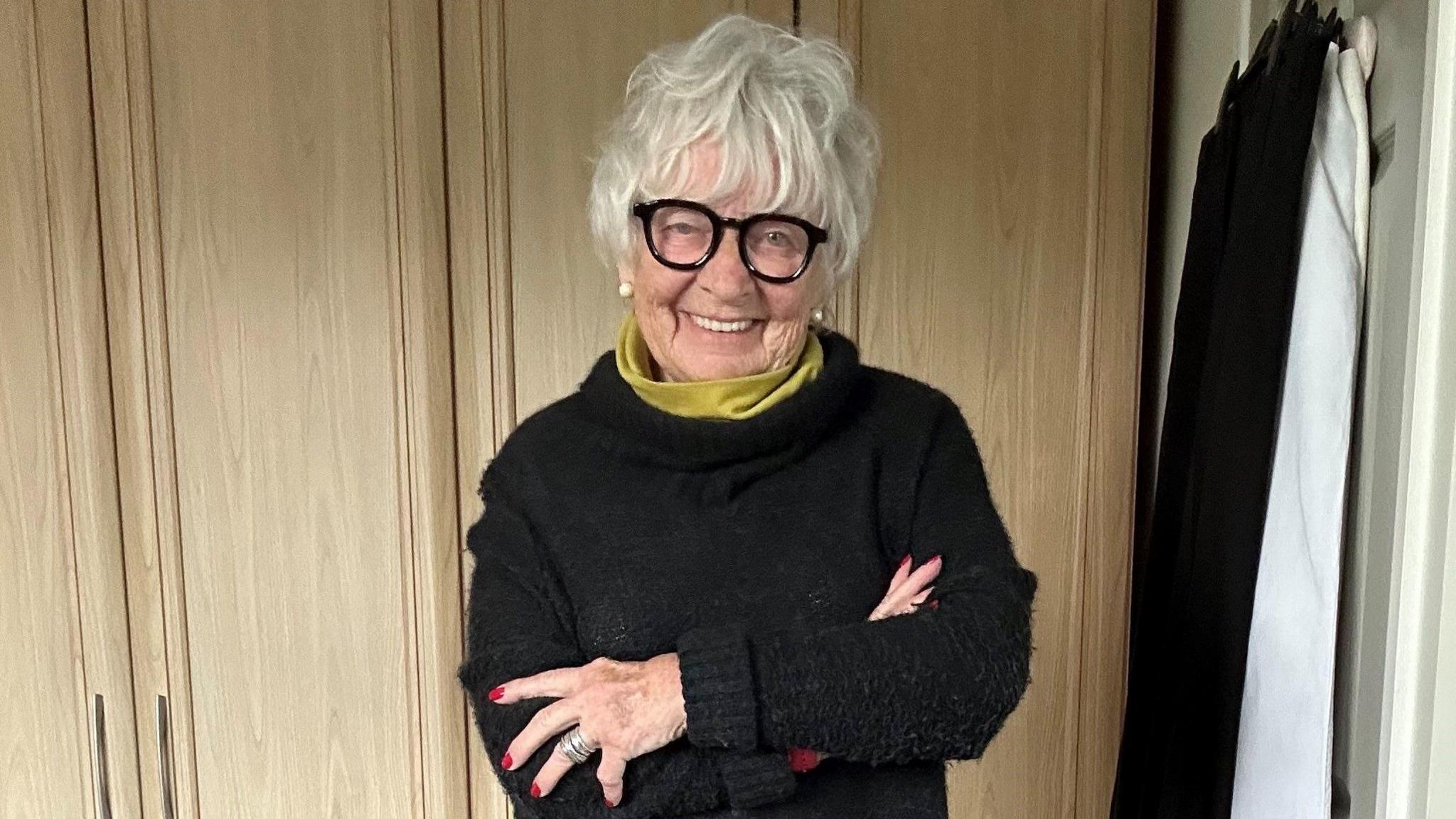 Mae Viv i'w gweld yma yn sefyll o flaen y camera. Mae ei breichiau wedi'u croesi. Mae hi'n gwisgo siwmper ddu gyda chris gwyrdd/melyn. Mae ganddi wallt gwyn byr ac mae'n gwisgo sbectol du.