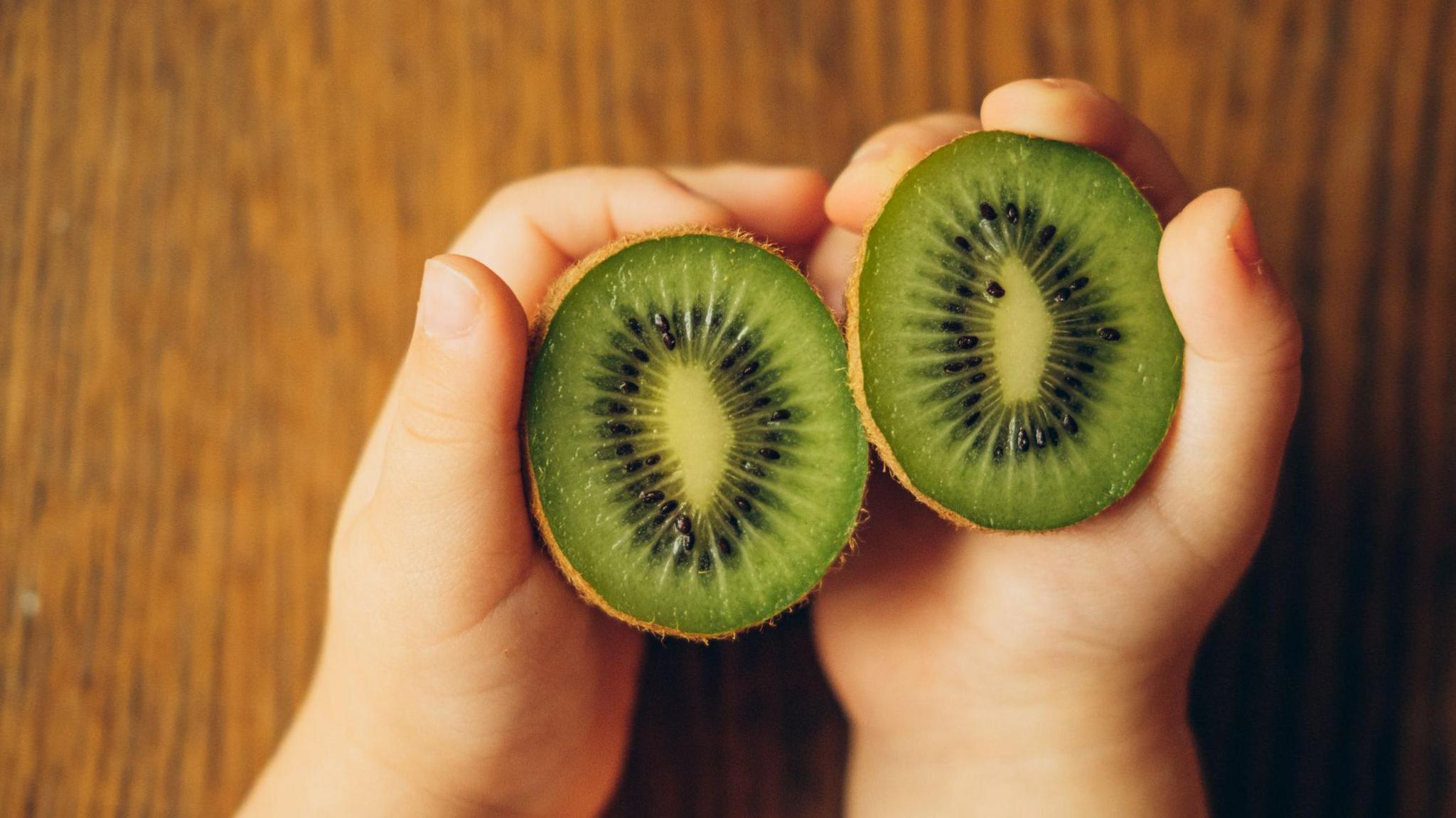 A child holds a kiwi, which has been cut into two halves, in their hands