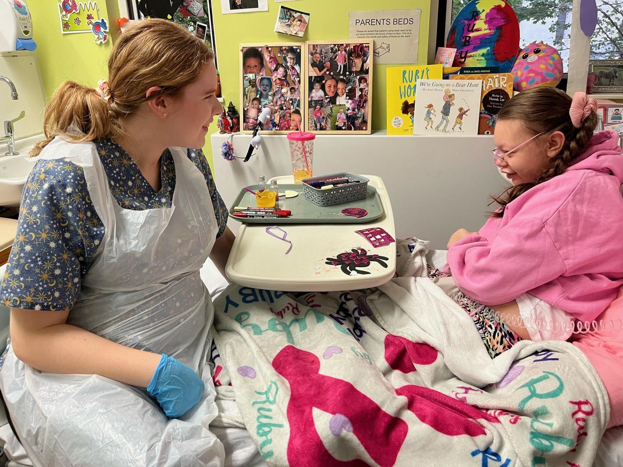 Patient Ruby, sitting up in her bed at Leeds Children's Hospital, and play specialist Megan Harpham