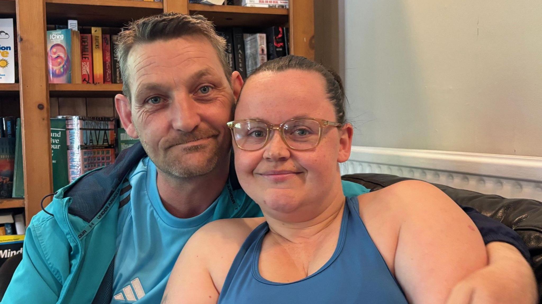 Mark and Elen take a selfie on a sofa with a bookcase full of books behind them. Elen has her hair tied back and is wearing glasses and a blue vest top, and Mark has short hair and stubble and is wearing a blue jacket and blue Adidas t-shirt.