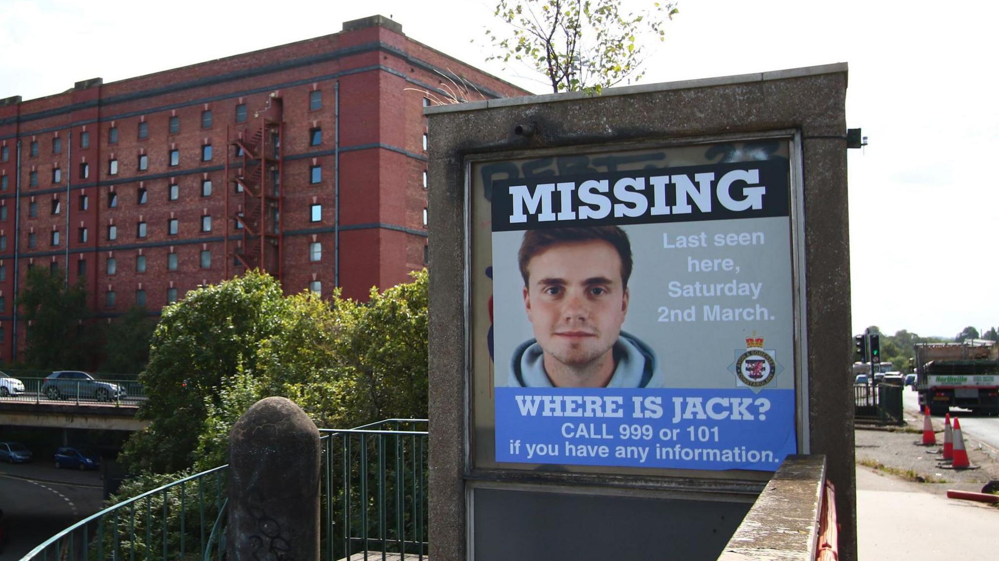 A poster is pictured close to the Cumberland Basin in Bristol. It has a picture of missing student Jack O'Sullivan in it and includes the working "where is Jack? Call 999 or 101 if you have any information". The photograph is taken on a sunny day and cars and one of the city's red brick former tobacco factories are visible in the background