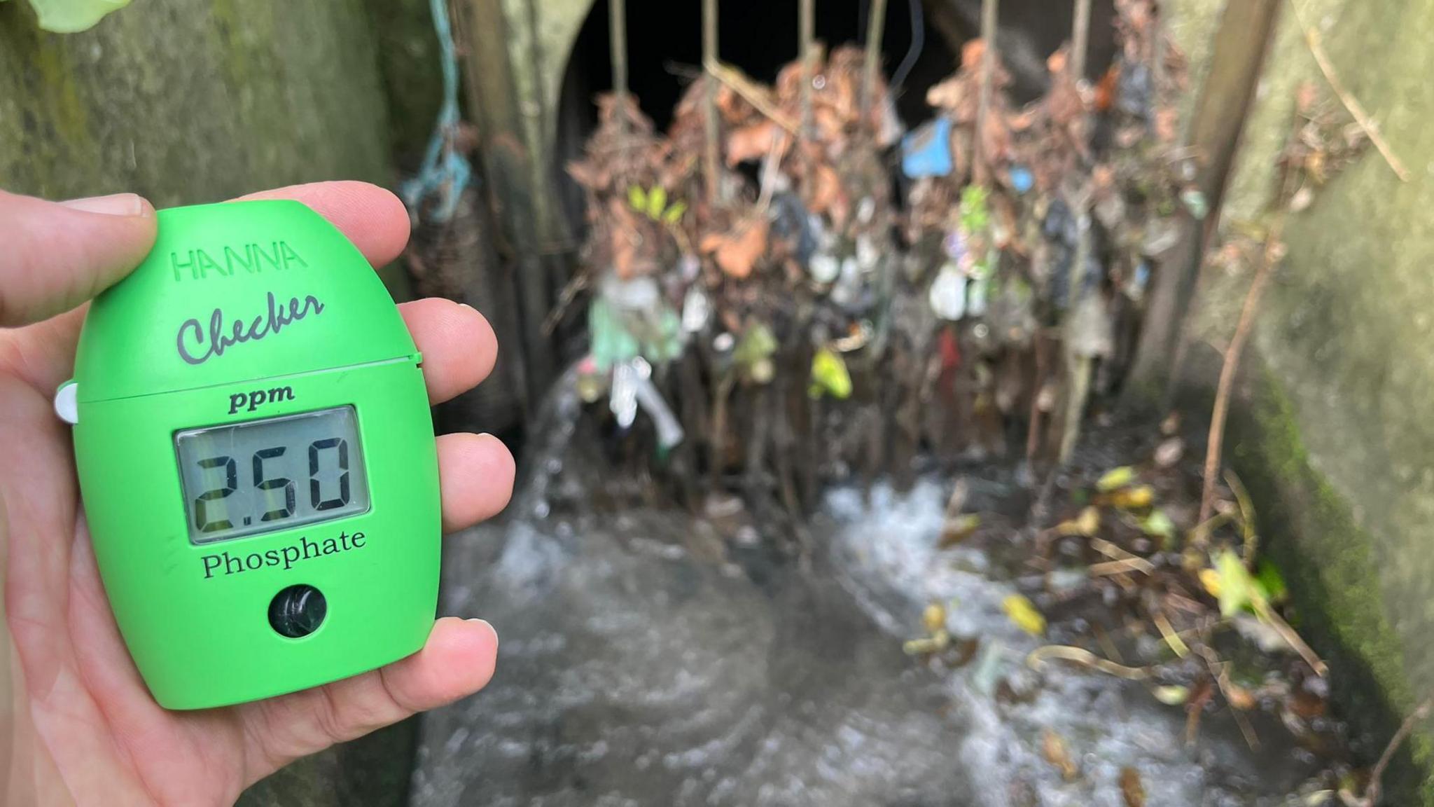 A hand holding a green phosphate reader in front of sewage outfall. The device shows a level of "2.50ppm".