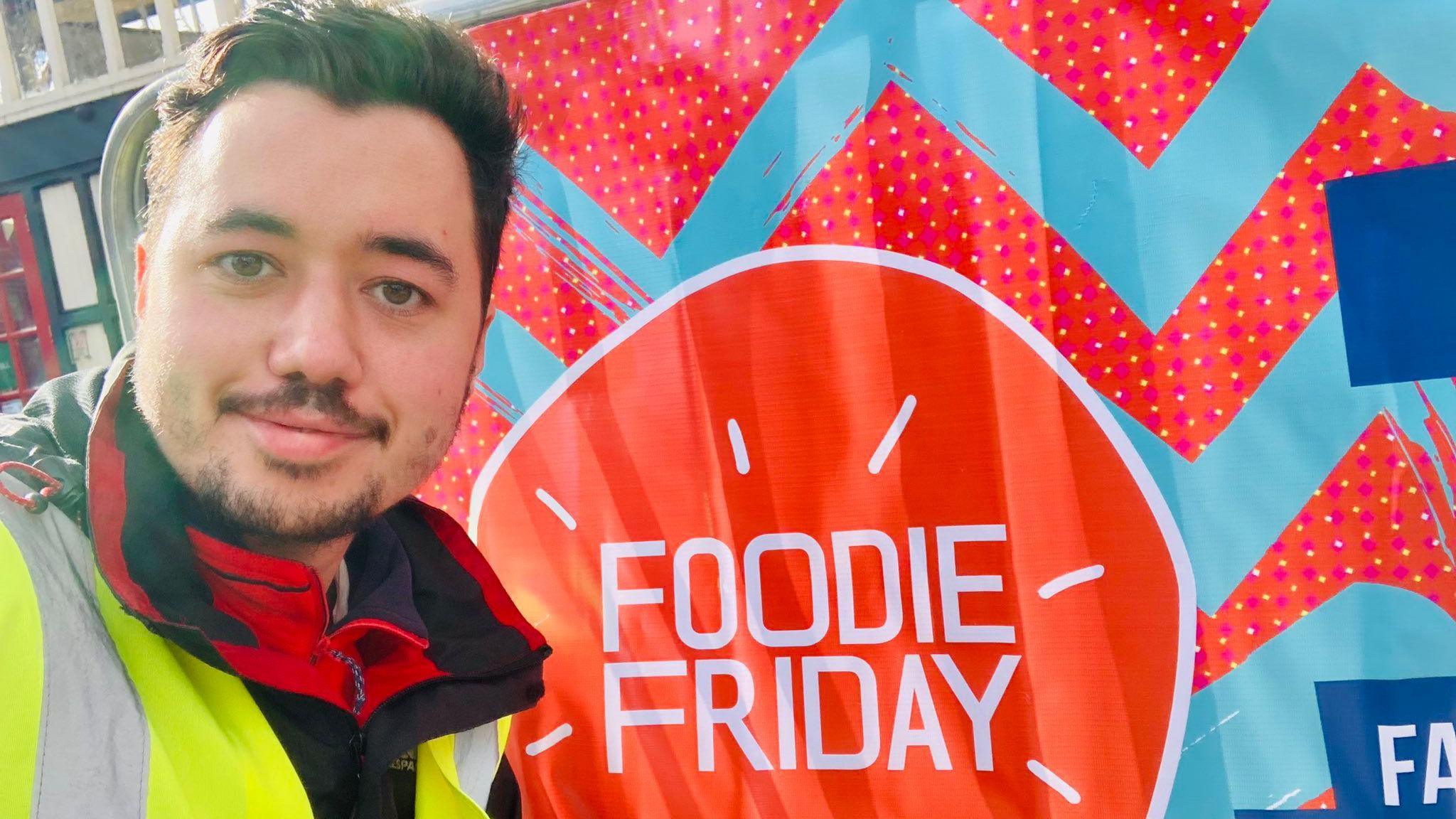Joe Barratt is standing in front of a colourful sign which advertises Foodie Friday. He is wearing a high-vis jacket and is smiling at the camera.