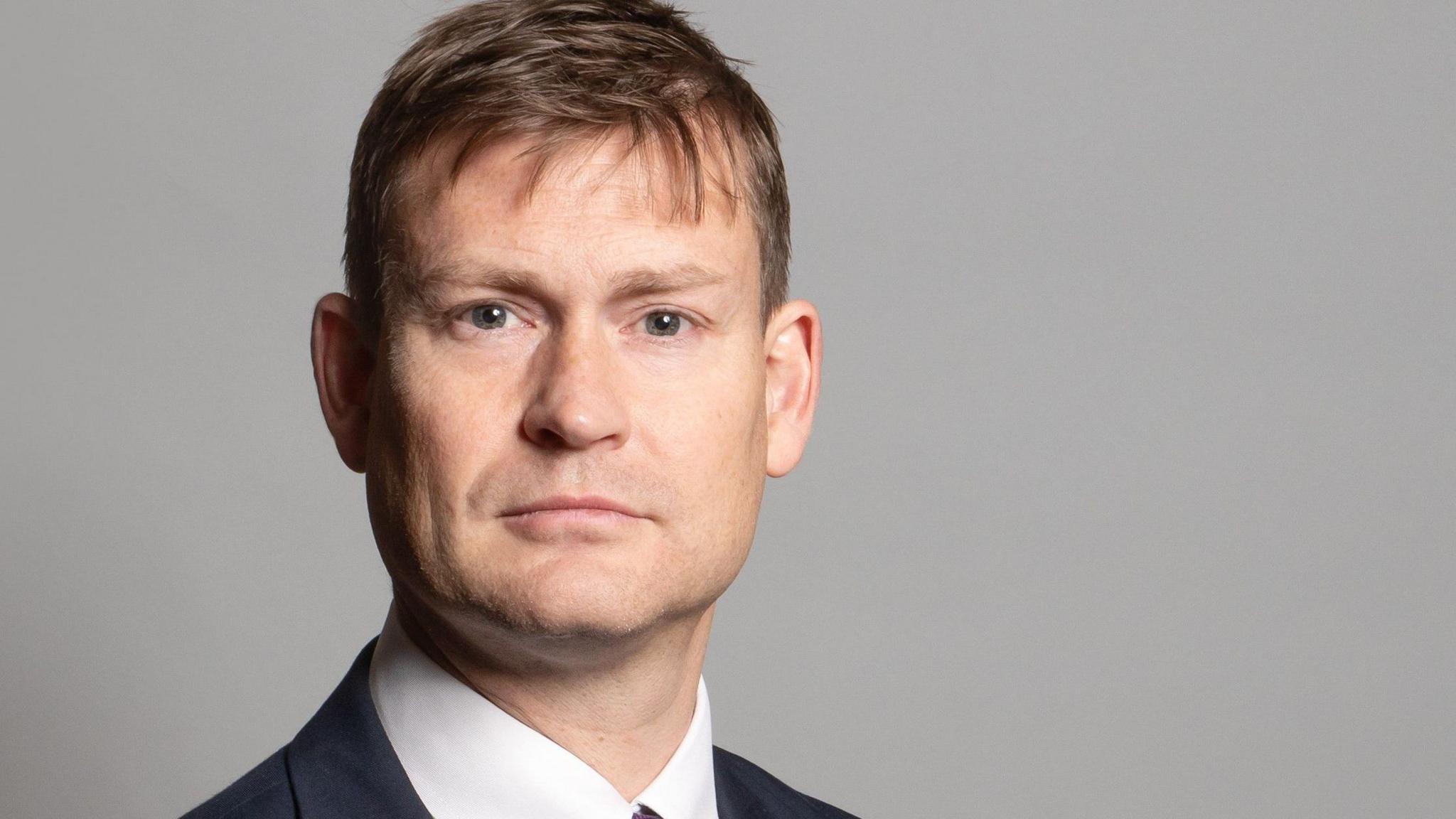 A parliamentary photo portrait of Mr Madders. He has short brown hair and is staring into the camera, and is wearing a dark suit, white shirt and purple tie. 