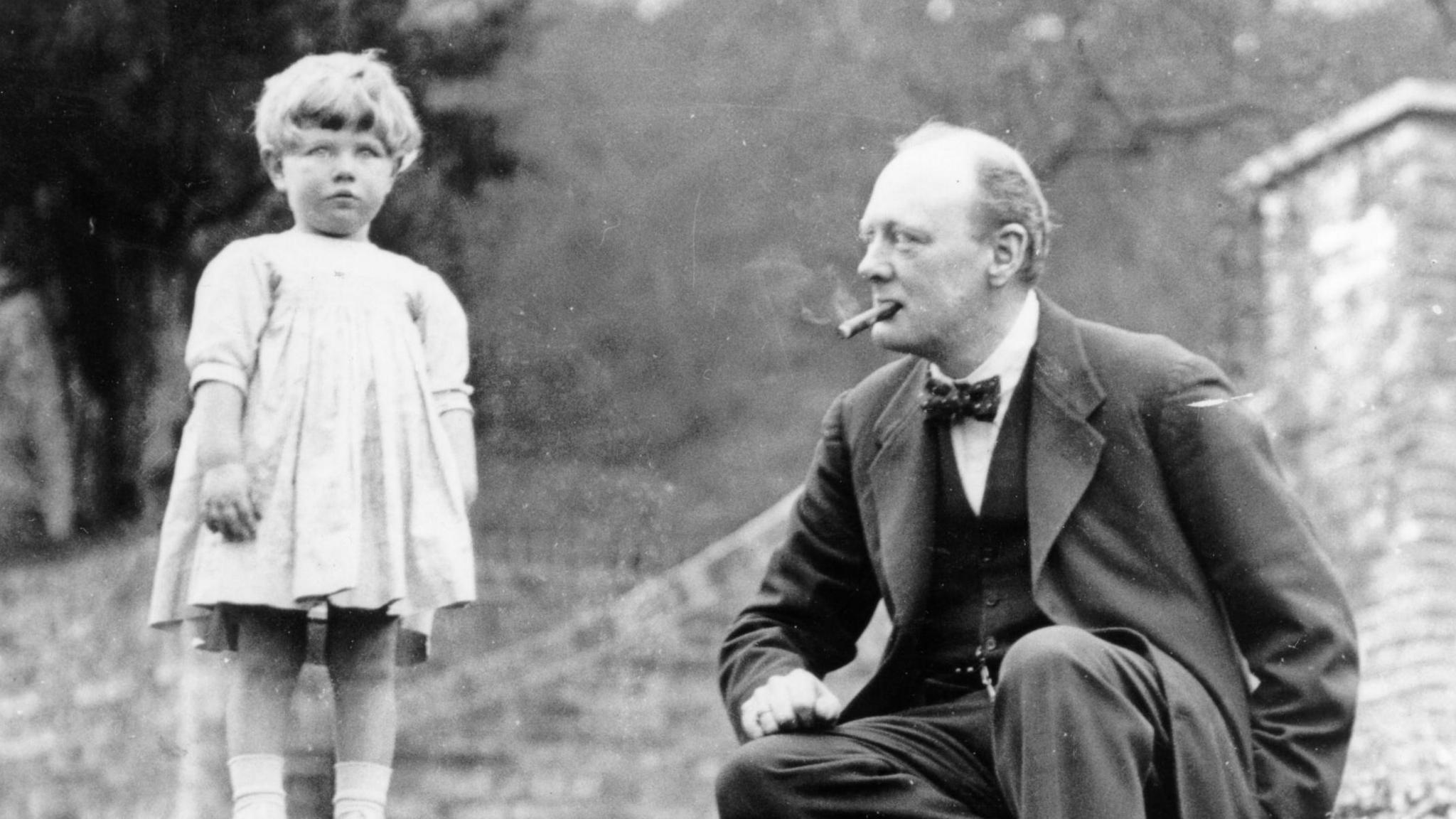 A black and white photograph of Winston Church in 1924, wearing a three piece suit and bow tie, smoking a cigar, sitting on garden steps, looking at his young daughter Mary, Chartwell, Kent