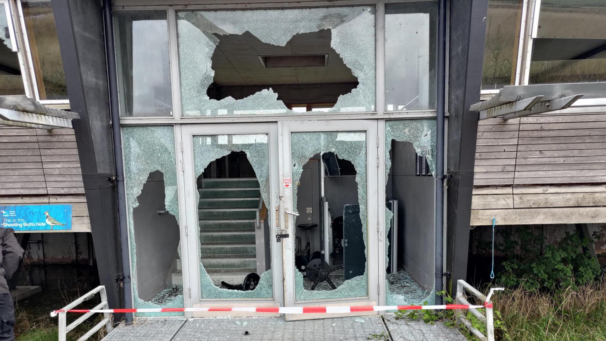A front entrance to a building with five completely smashed windows and red tape across the front.